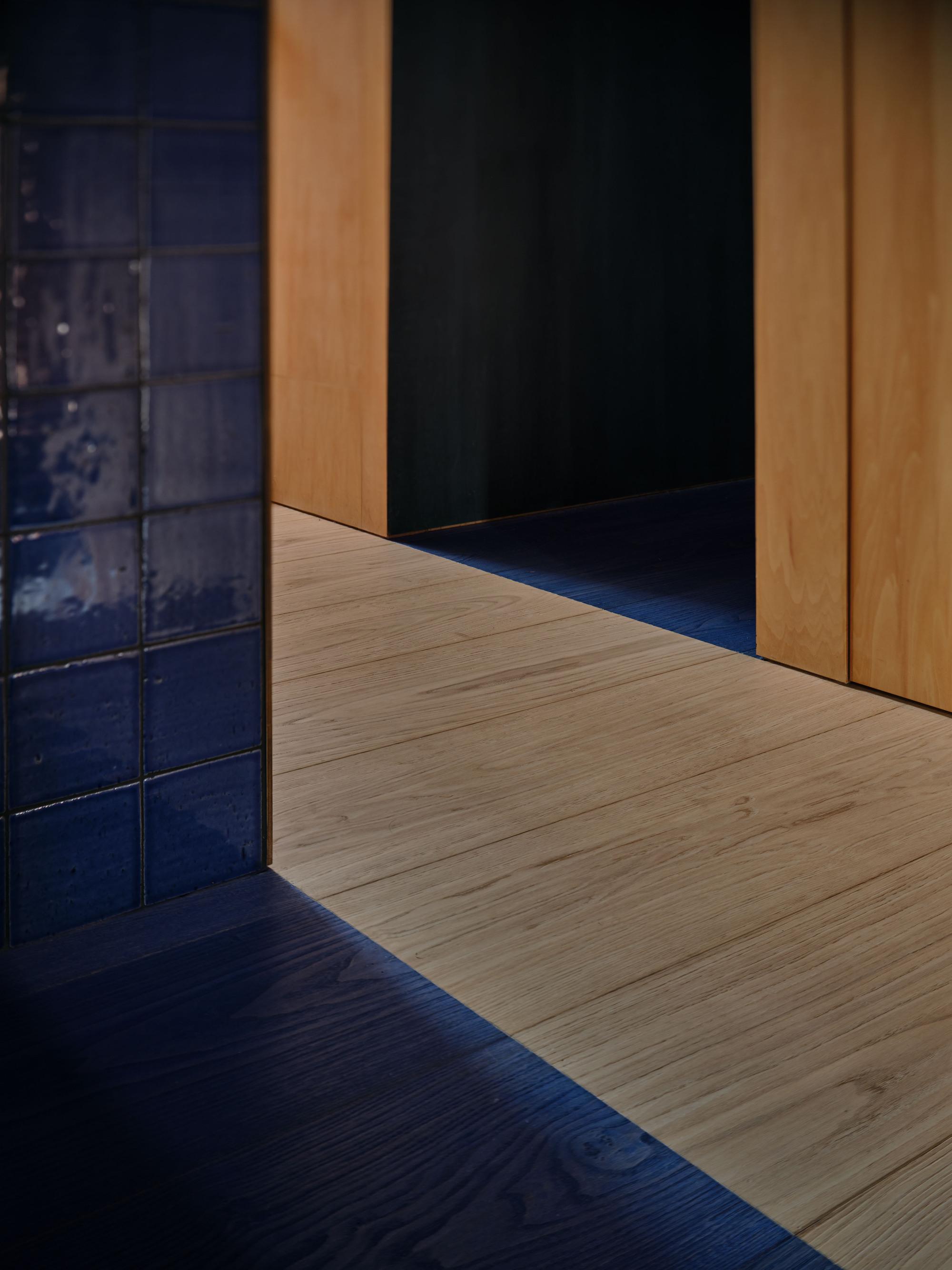Threshold where glazed blue tile and dark blue floorboards meet pale timber flooring and plywood panels in low light