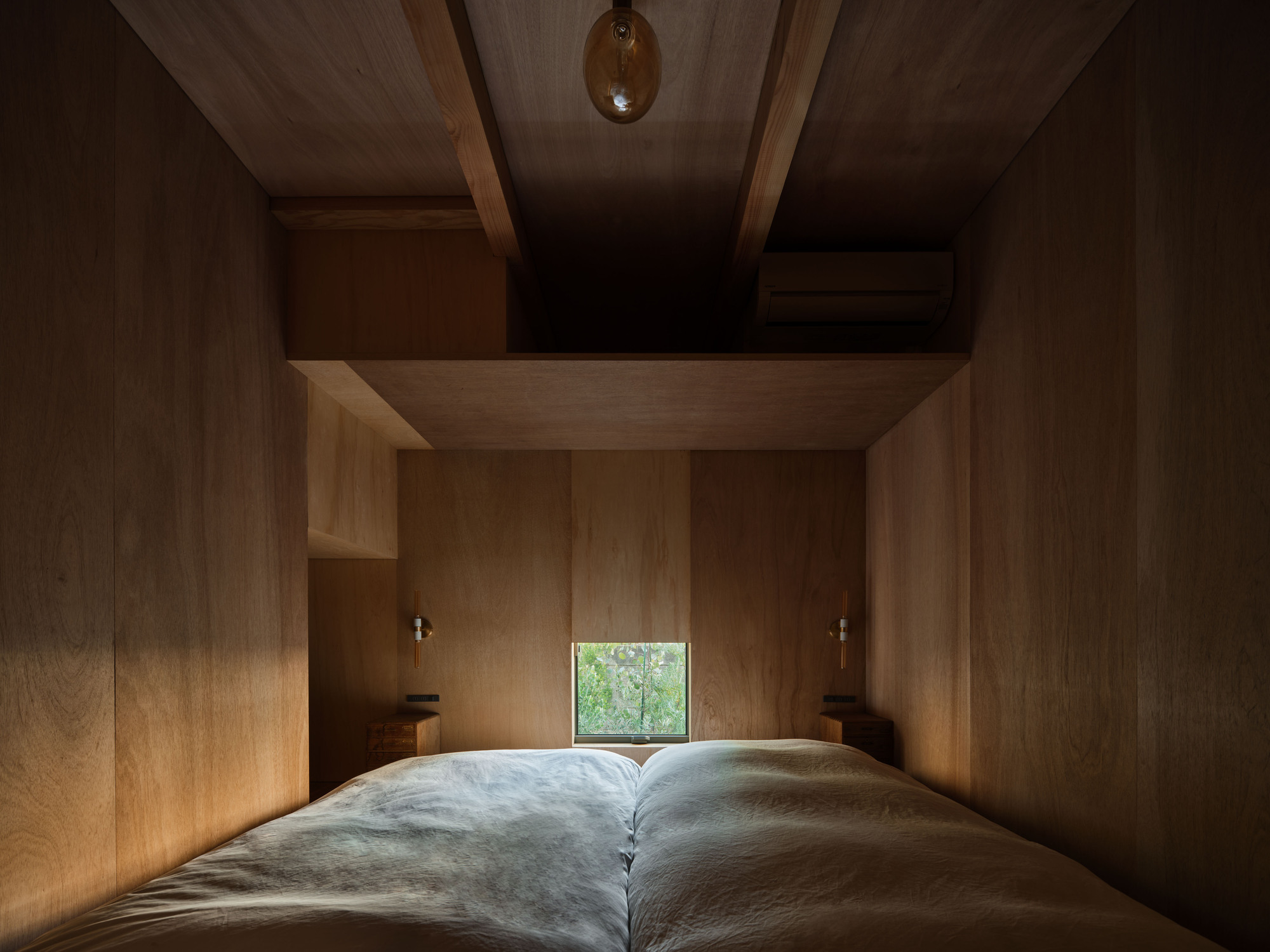 Plywood-lined bedroom with exposed ceiling beams and a small square window glowing green with foliage behind the bed