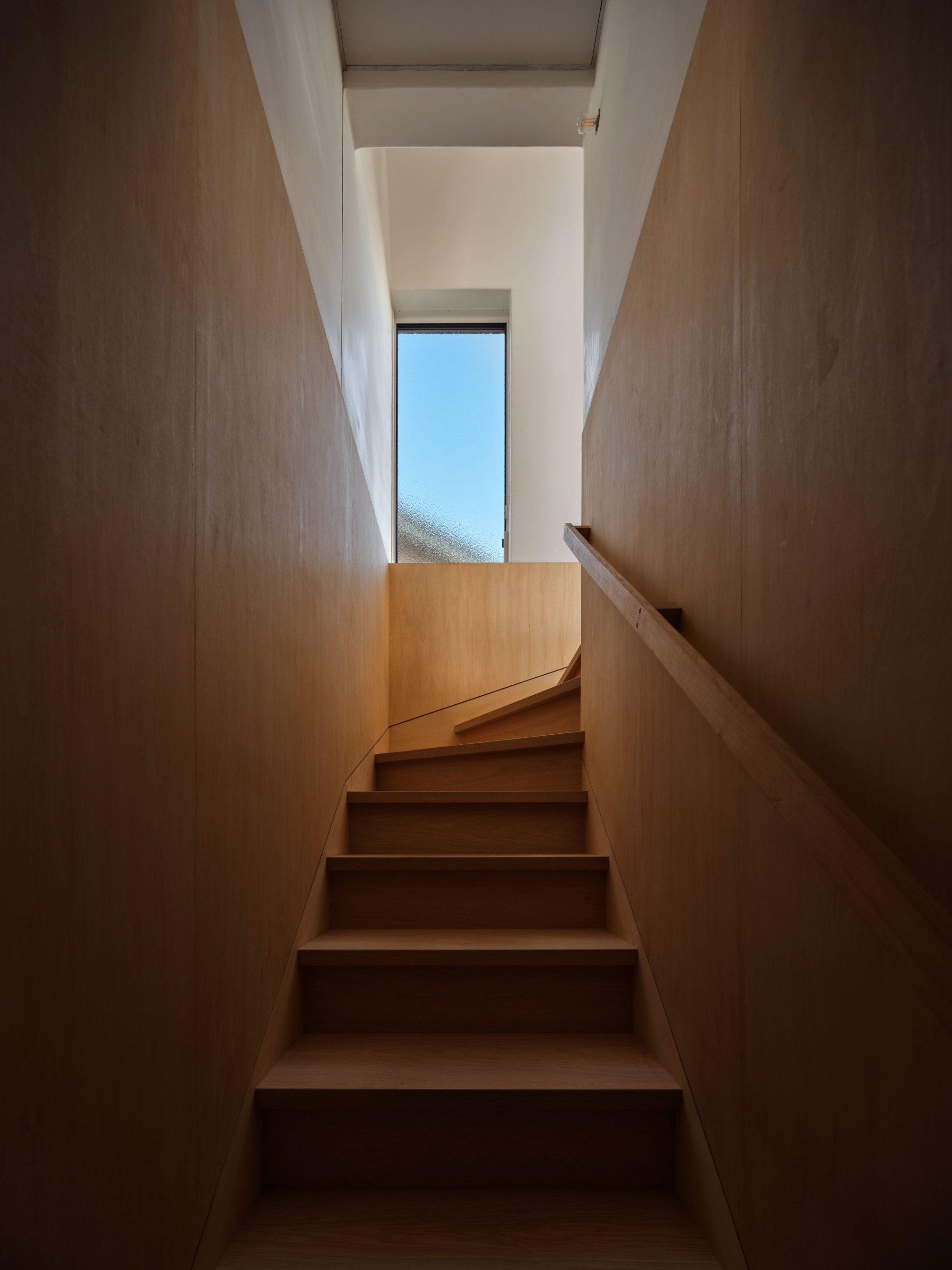 Narrow timber staircase with a wooden handrail rising toward a tall window filled with clear blue sky