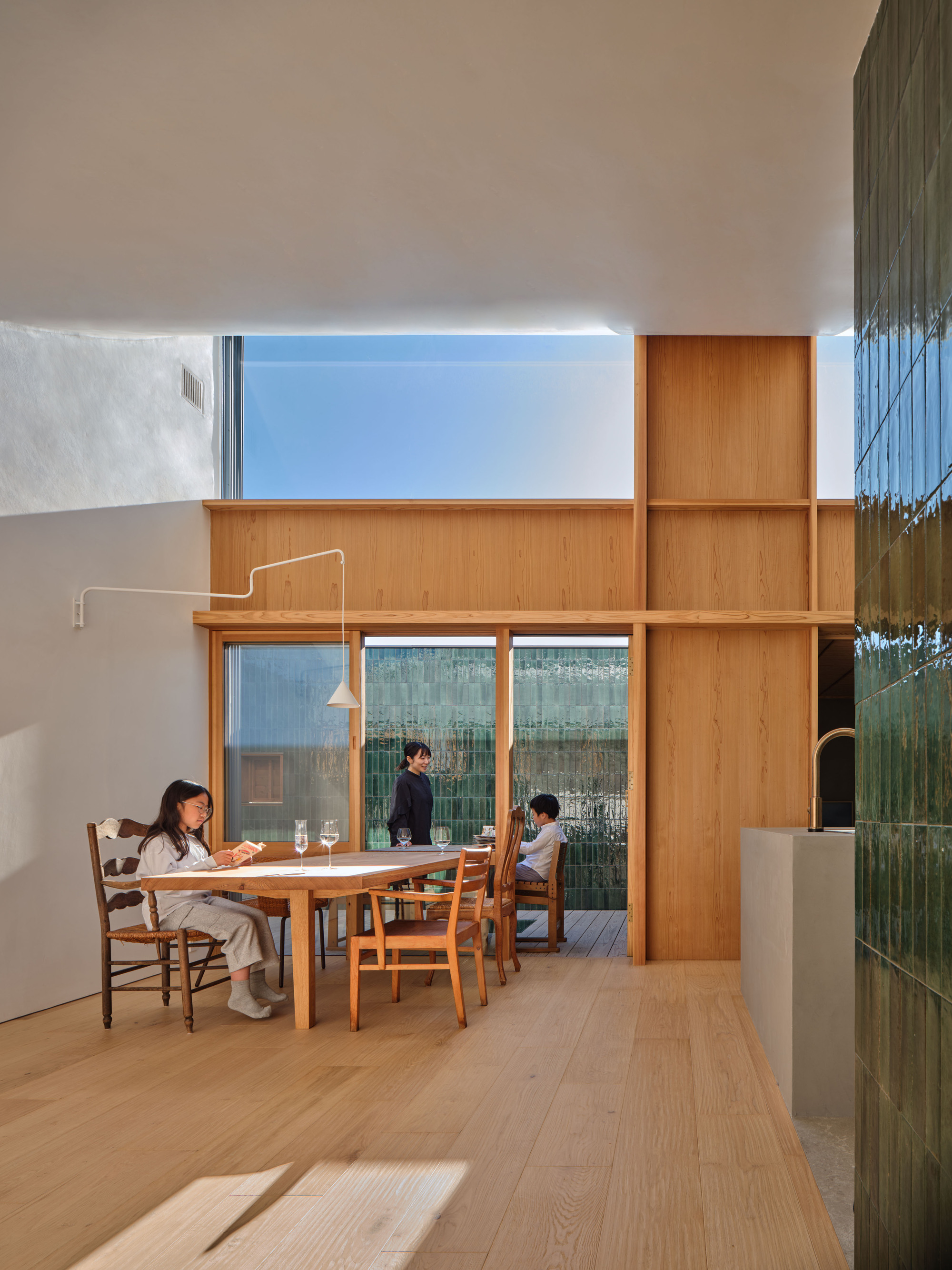 Dining area with a timber table, a girl reading and a woman standing at sliding glass doors facing a green tile wall