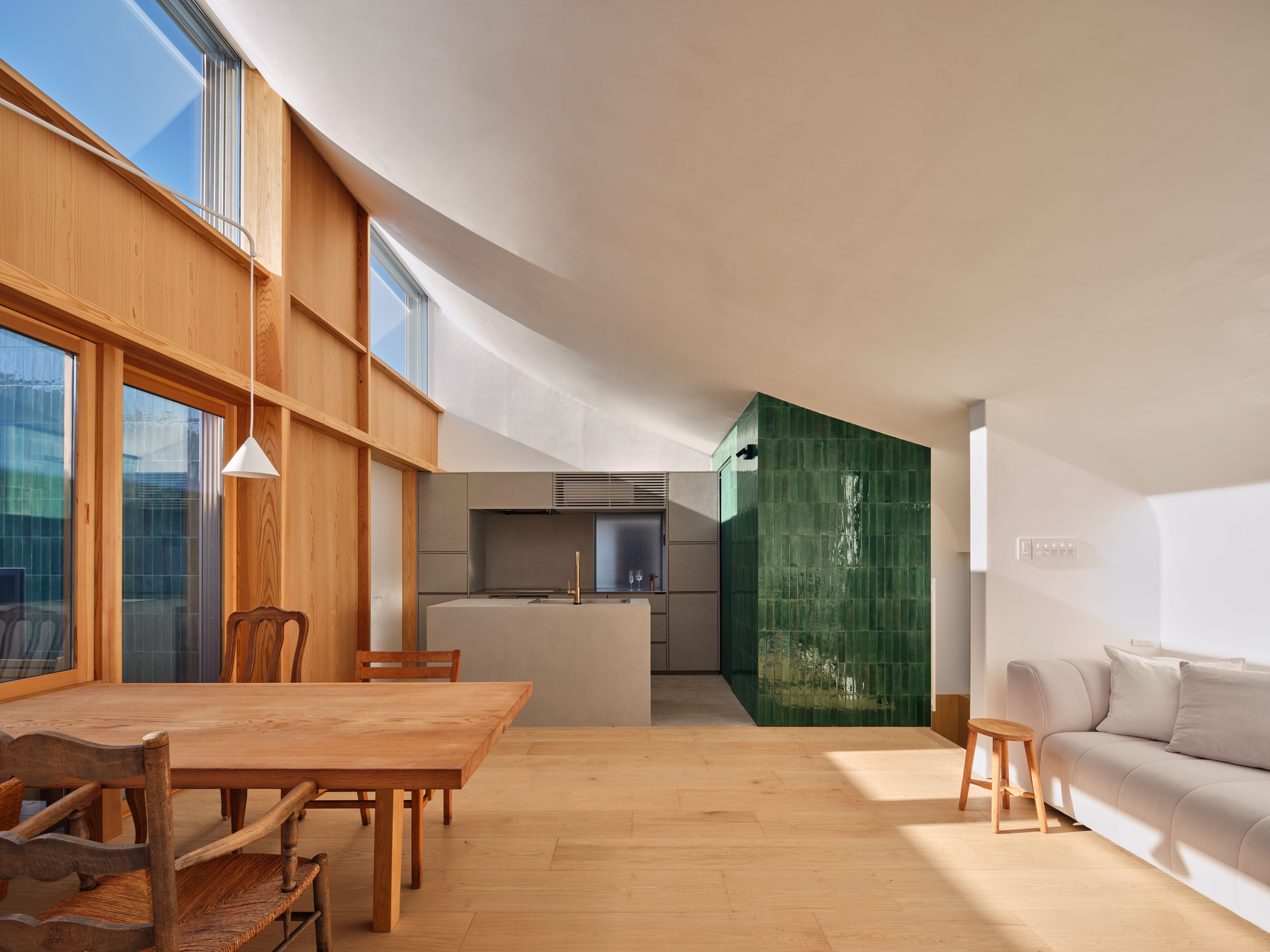 Open living space with a green glazed tile volume, stone kitchen island and timber dining table under a curved plaster ceiling