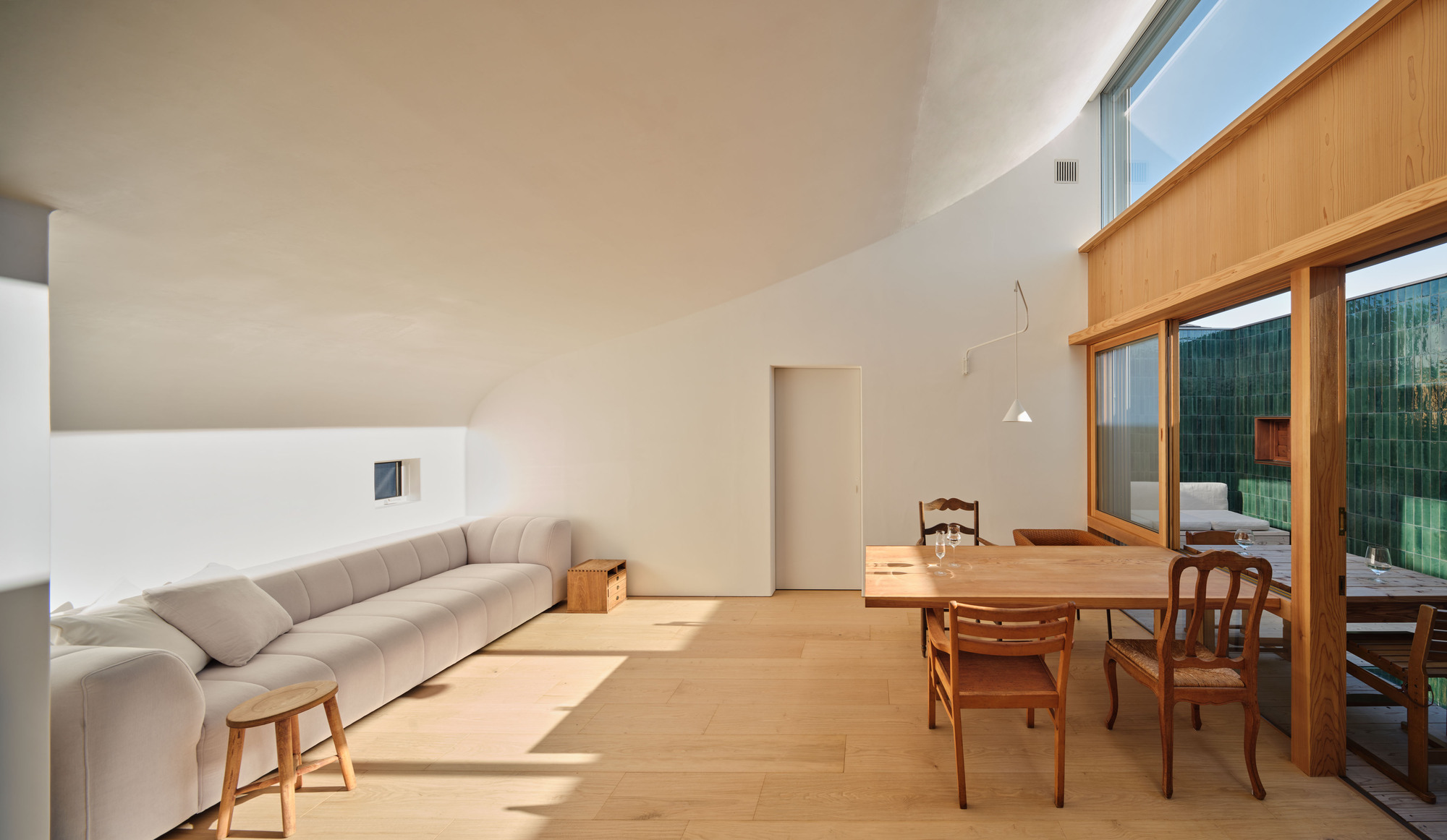 Long view of the grey sofa and dining table beneath a curving white ceiling, with sunlight falling across the oak floor