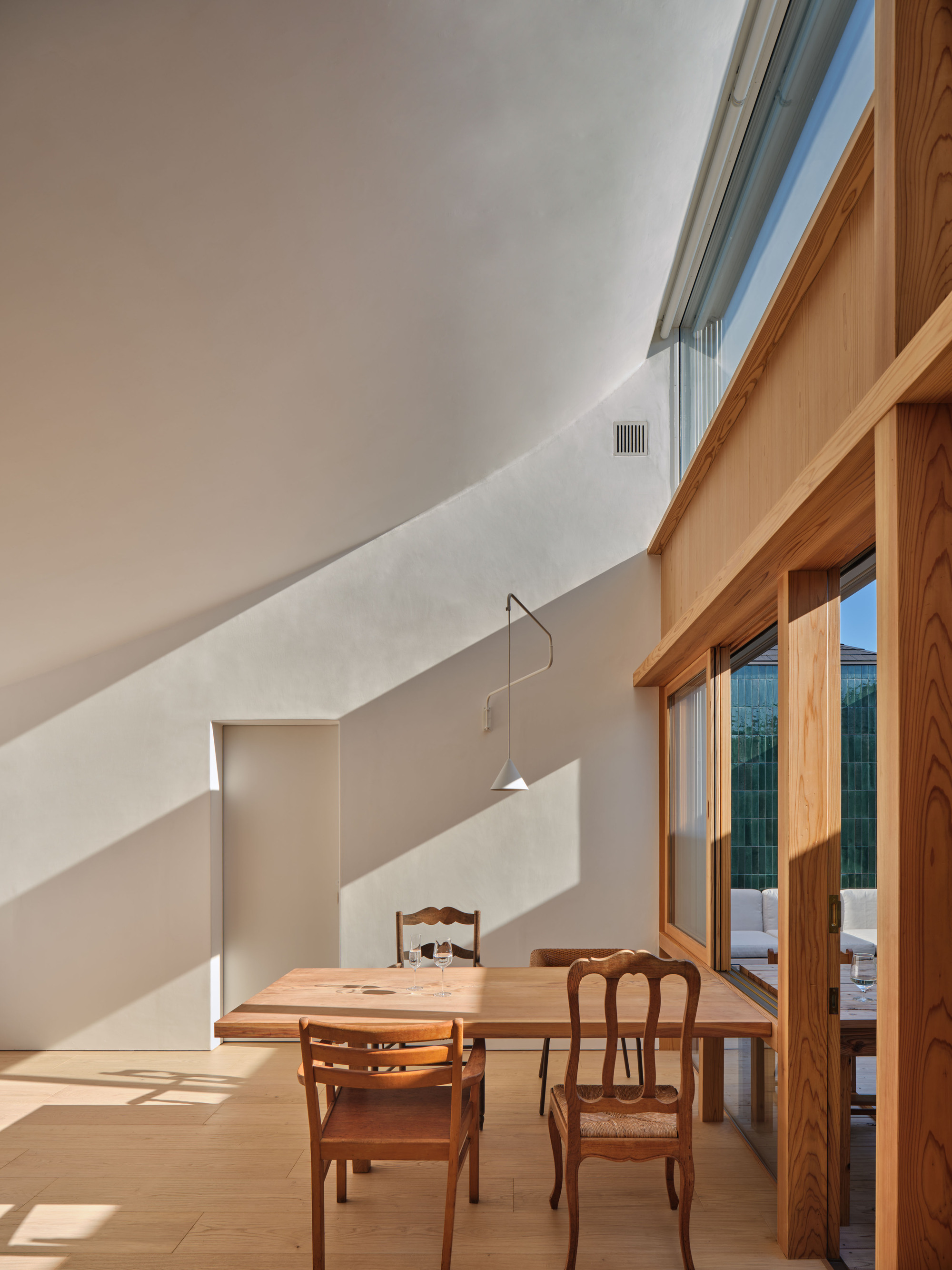 Dining table and spindle chairs beside a timber window wall, with afternoon sun cutting diagonal shadows across the curved plaster wall