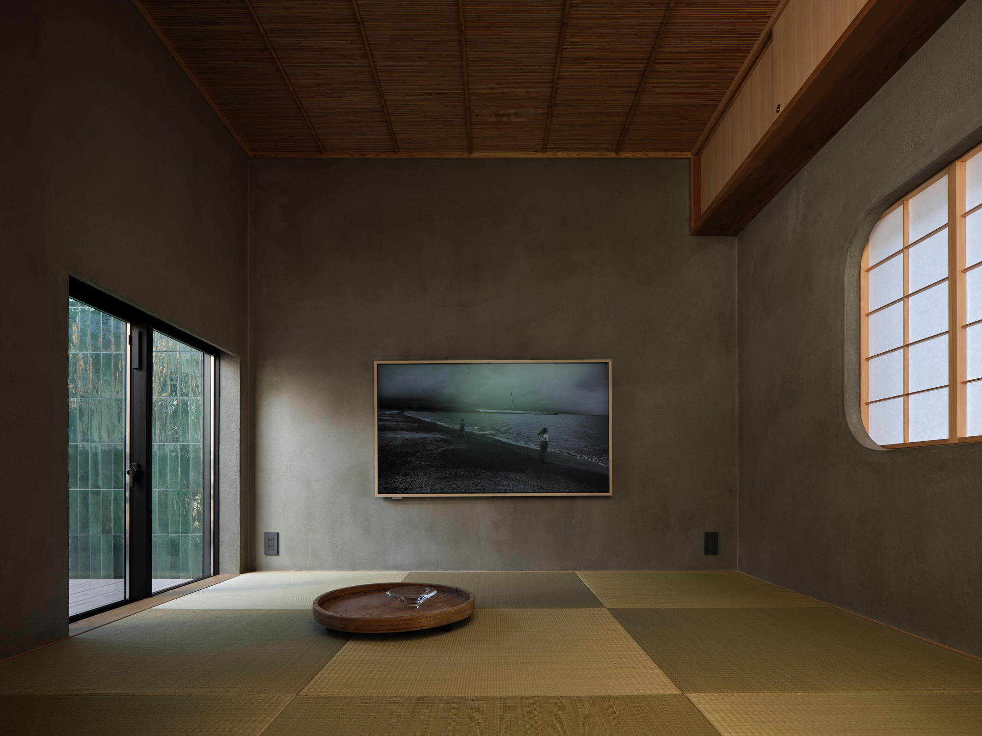 Tatami room with grey plaster walls, a framed seascape photograph, a round wooden bowl on the mats and a rounded shoji window