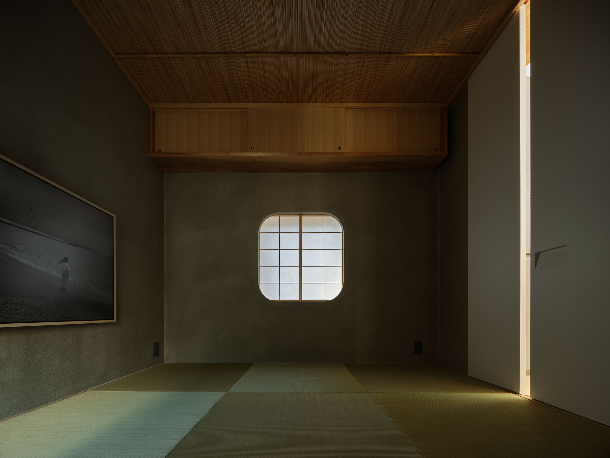 Tatami room seen toward a small rounded shoji window, with a bamboo ceiling, built-in timber cabinet and a thin slit of light at the door