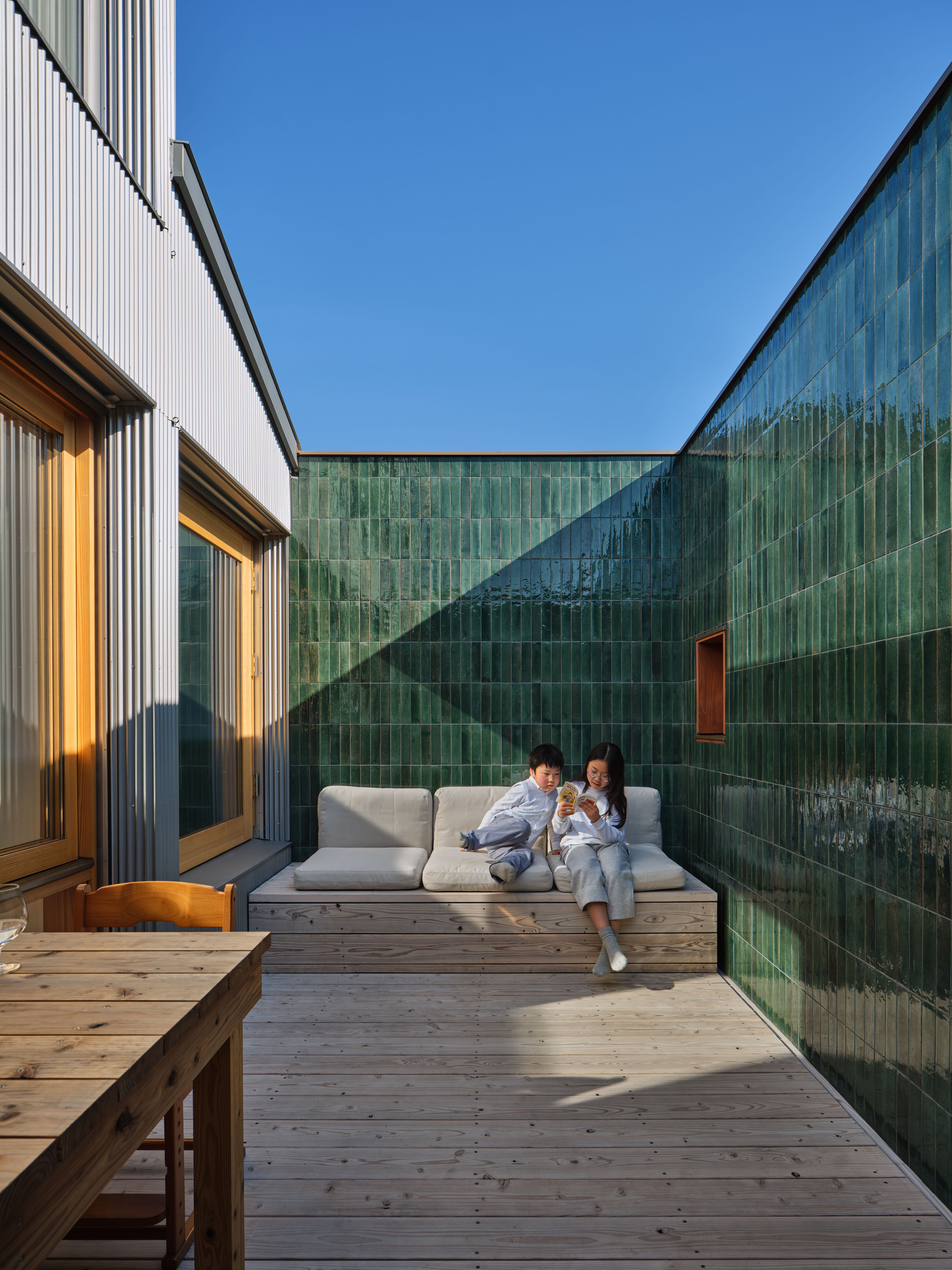 Two children reading on a built-in cushioned bench against green glazed tile, with a shadow cutting across the deck