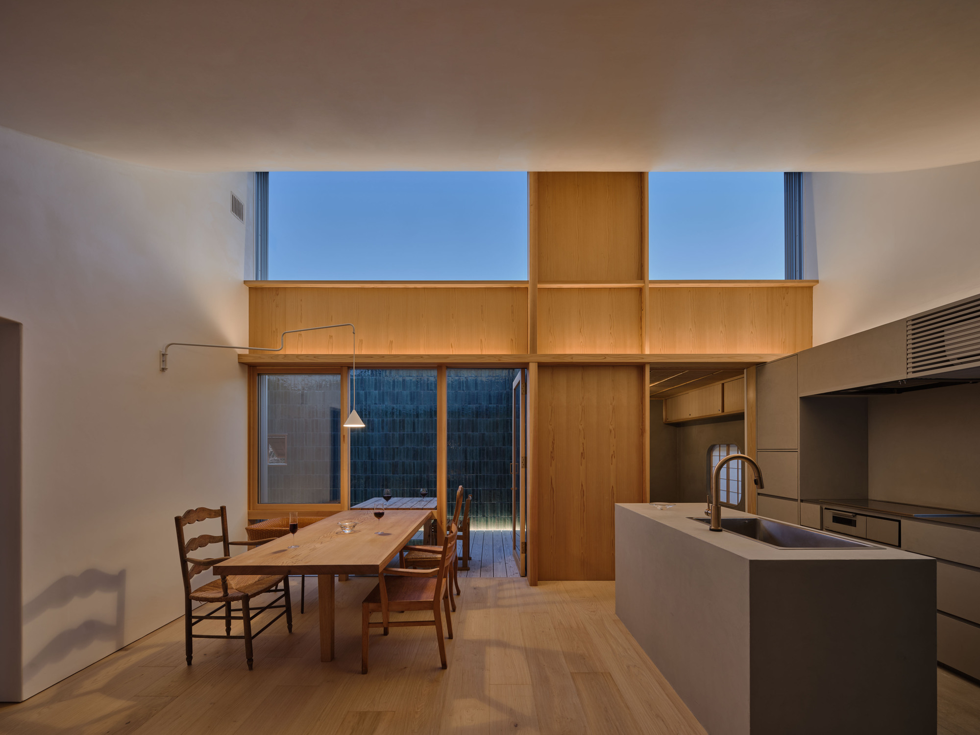 Open kitchen and dining space with a timber clerestory wall and concrete island, lit by blue dusk light