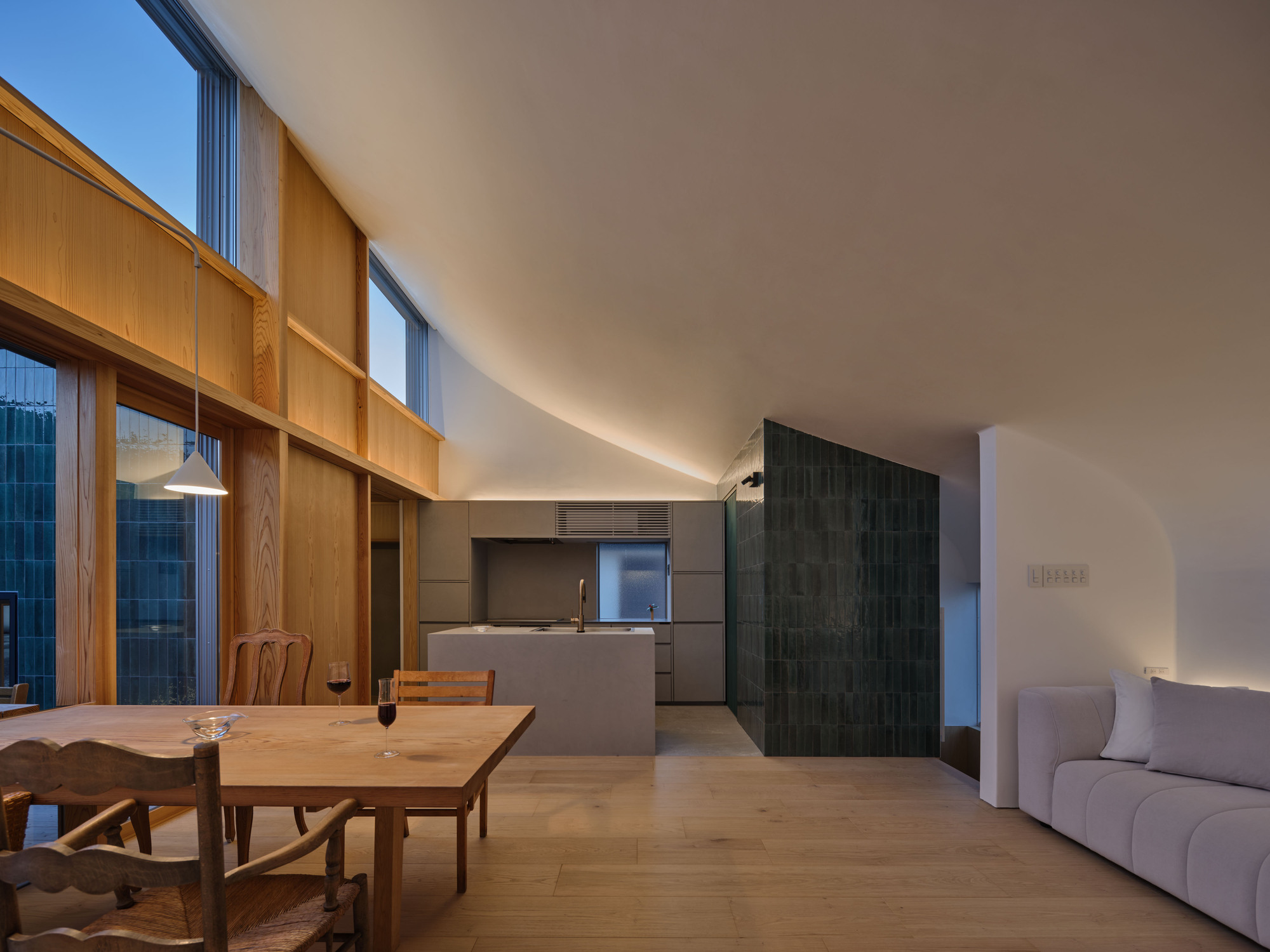 Living area under a curved white ceiling, with a green tile volume, kitchen island and sofa at dusk