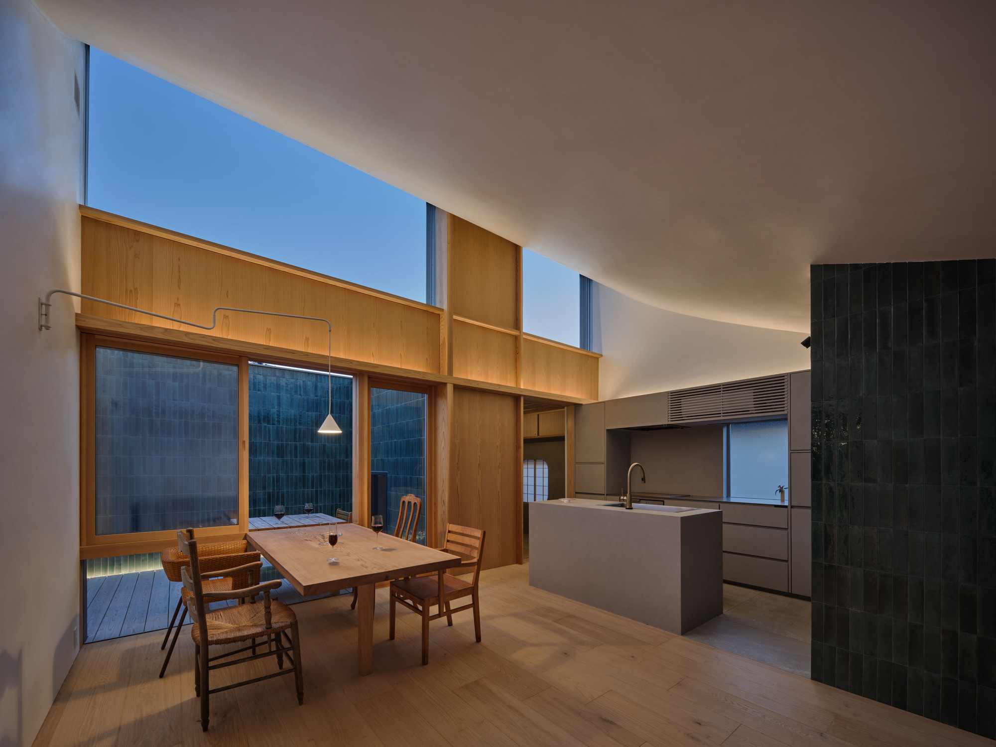 Dining table and stone-topped kitchen island beneath a curved white ceiling, with a dark green tile wall at dusk