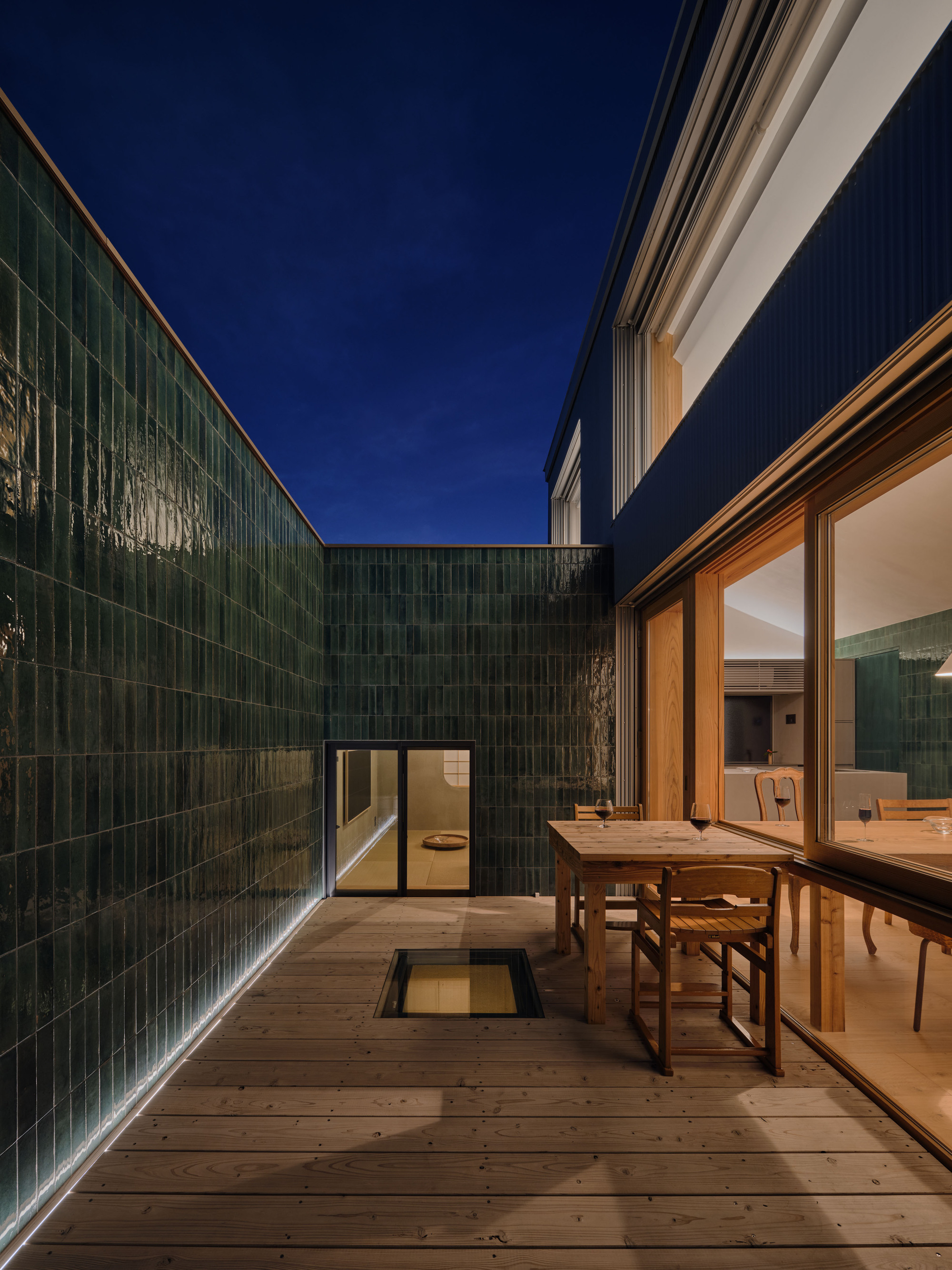 Timber deck courtyard enclosed by glossy green tile walls, with a dining table by the glass doors under a deep blue night sky