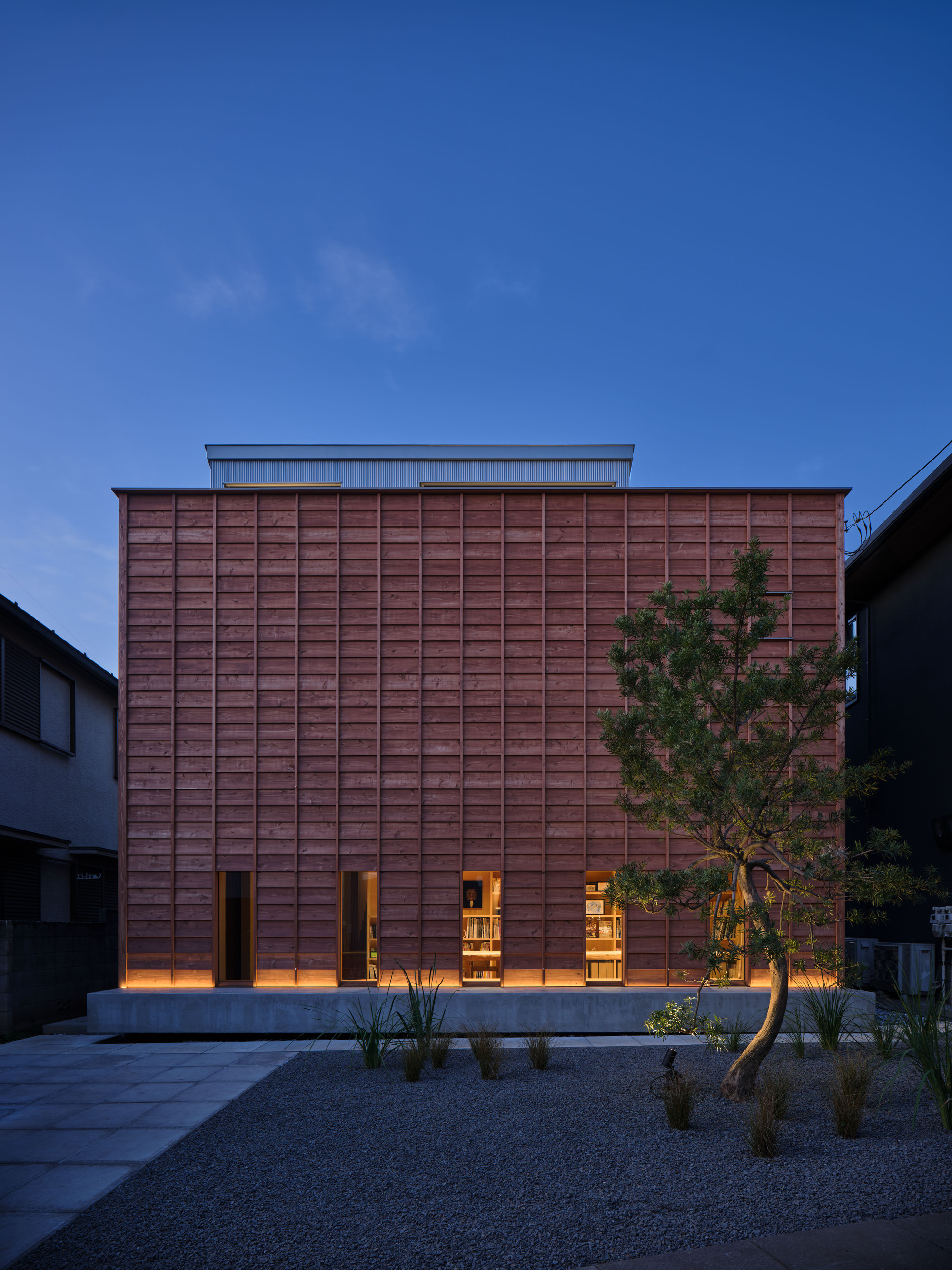Street facade clad in red-brown timber boards with lit ground-floor openings, a twisted pine tree and gravel garden at dusk