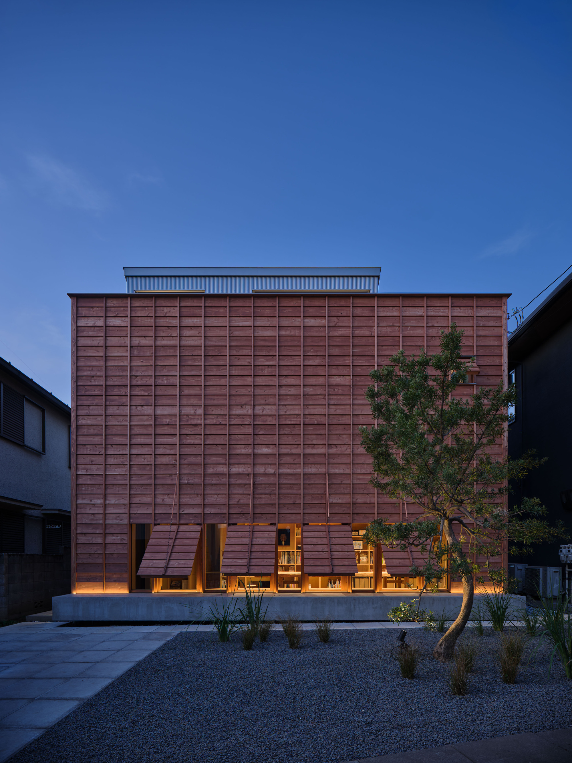 Same timber facade with hinged wooden shutters propped open along the ground floor, a pine tree and gravel in the blue dusk