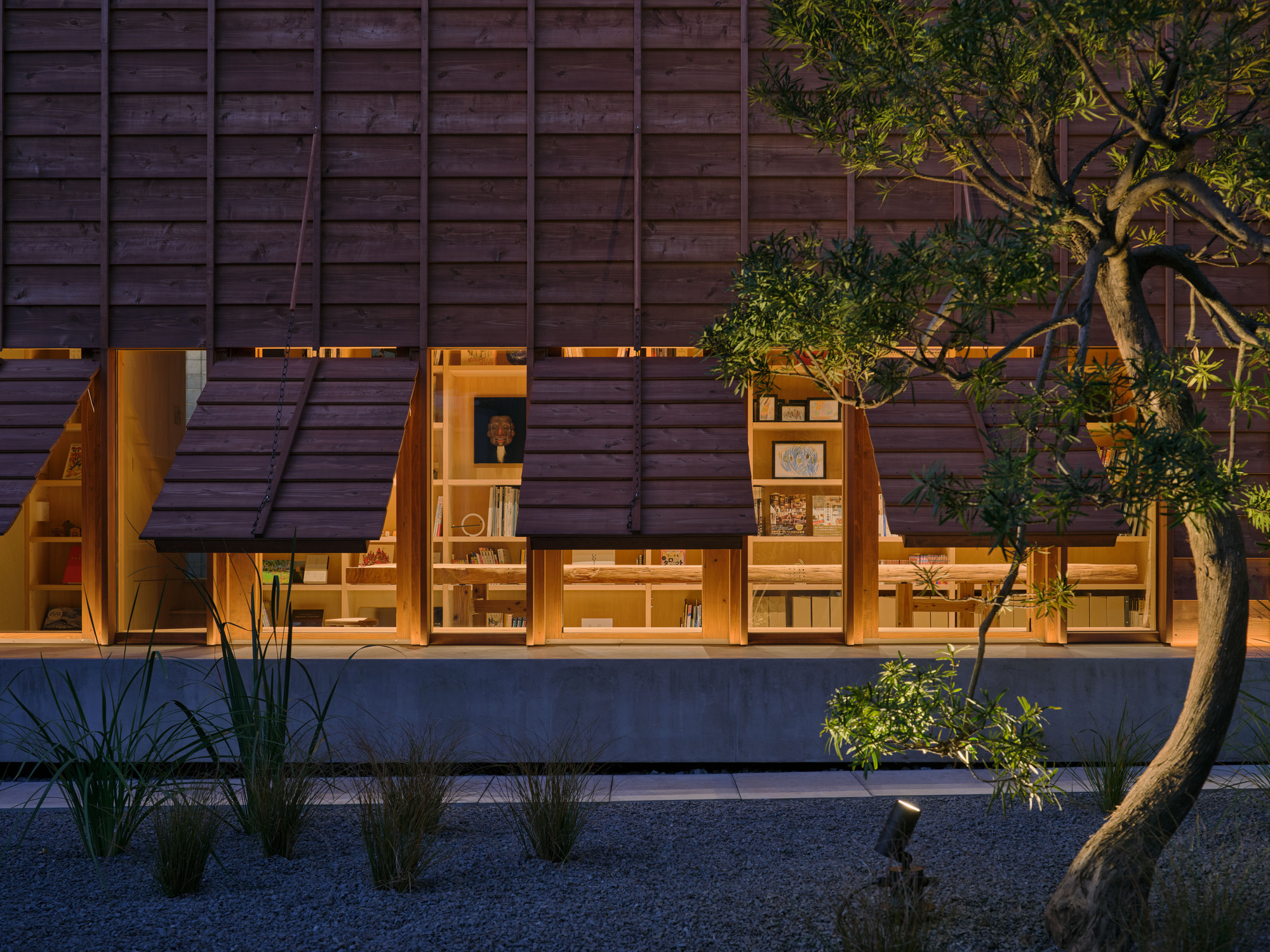 Close view of open shutters above a library window wall lit from within, with a pine branch and a ground spotlight in the gravel