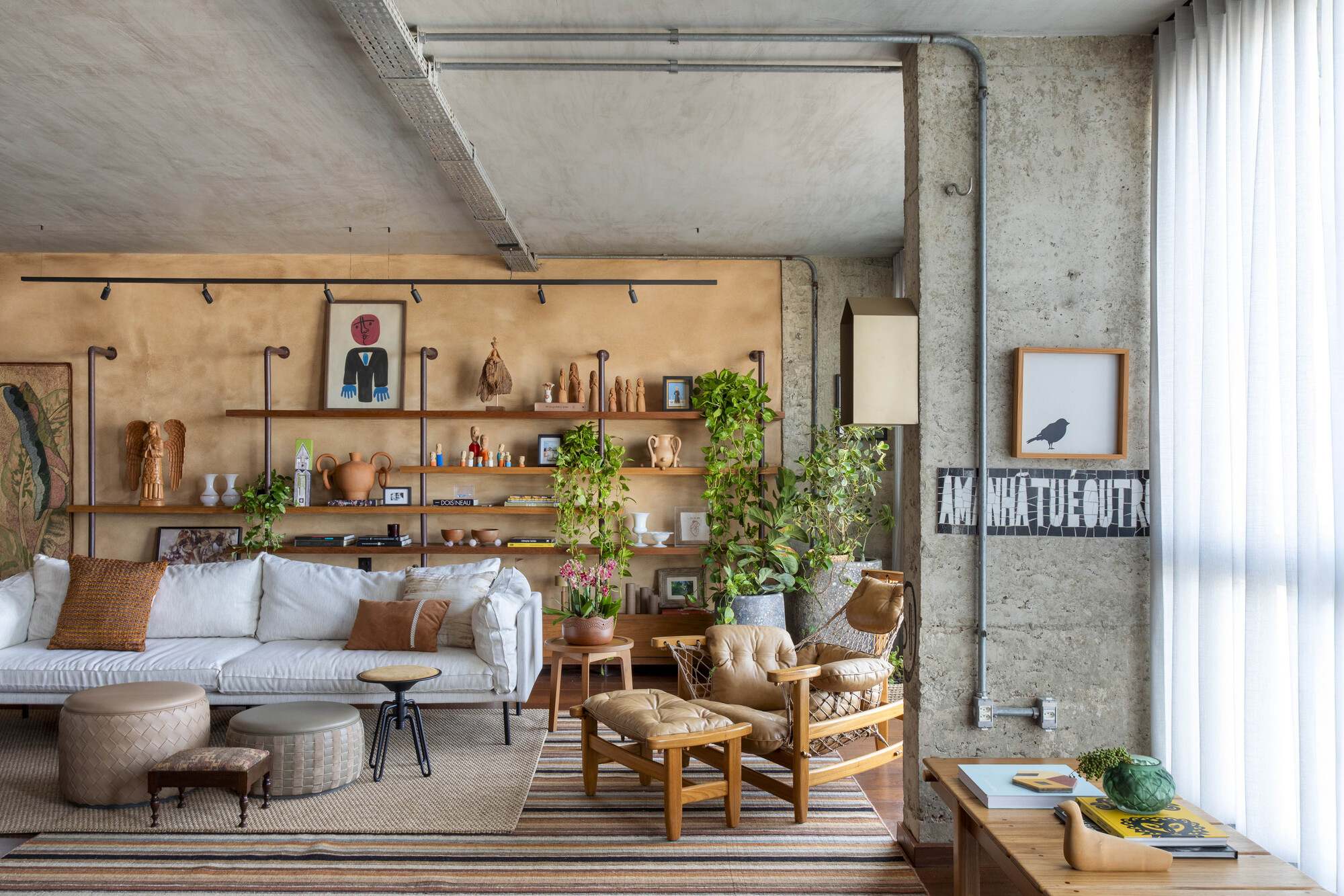 Living room with white sofa, leather lounge chair and open shelves of clay figures against an ochre wall, lit by sheer-curtained daylight