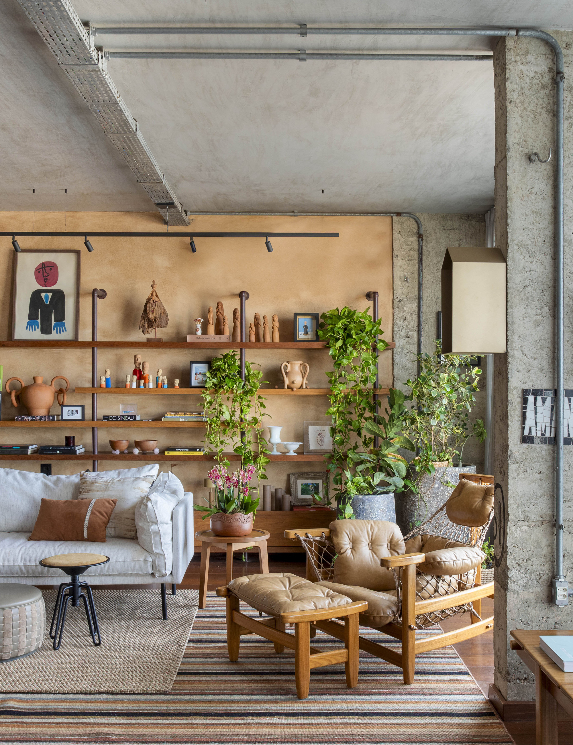 Wood-framed lounge chair and ottoman on a striped rug beside trailing plants and shelves of terracotta objects under a concrete ceiling