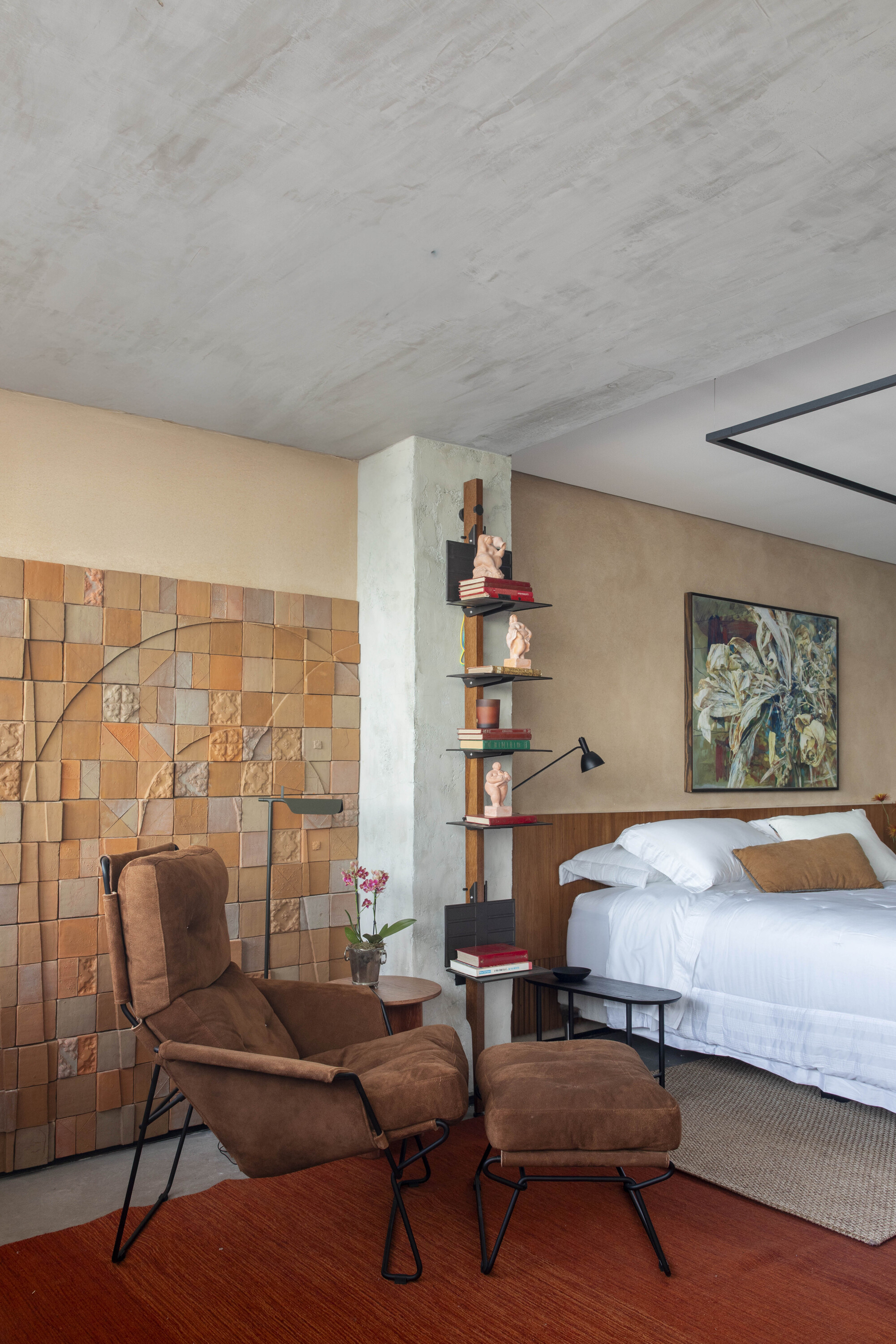 Bedroom corner with a brown suede armchair before a wall of handmade terracotta tiles, a slim shelf and a bed in soft daylight