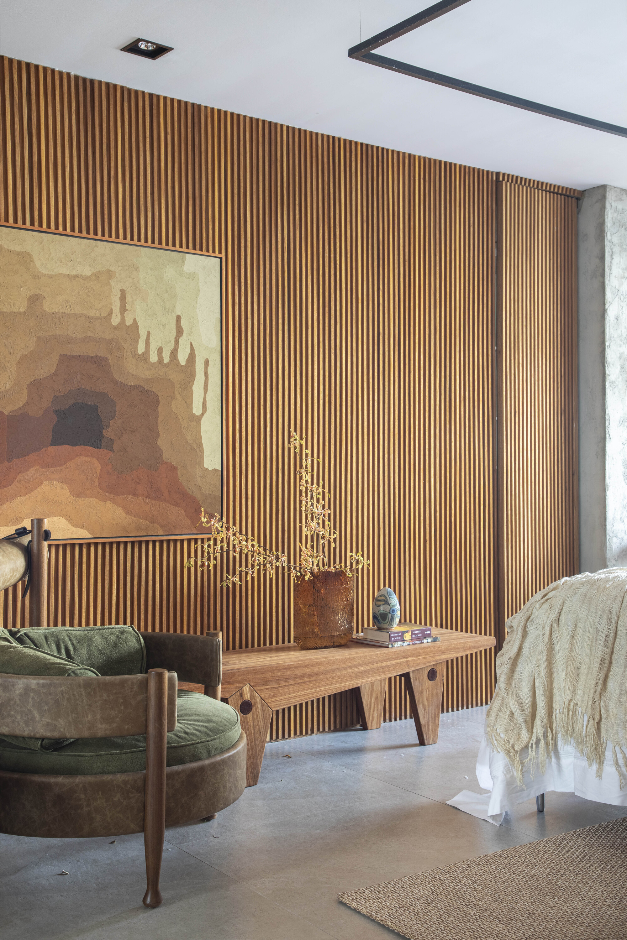 Vertical timber slat wall with a large abstract landscape painting, a wooden bench, dried branches and a green velvet armchair