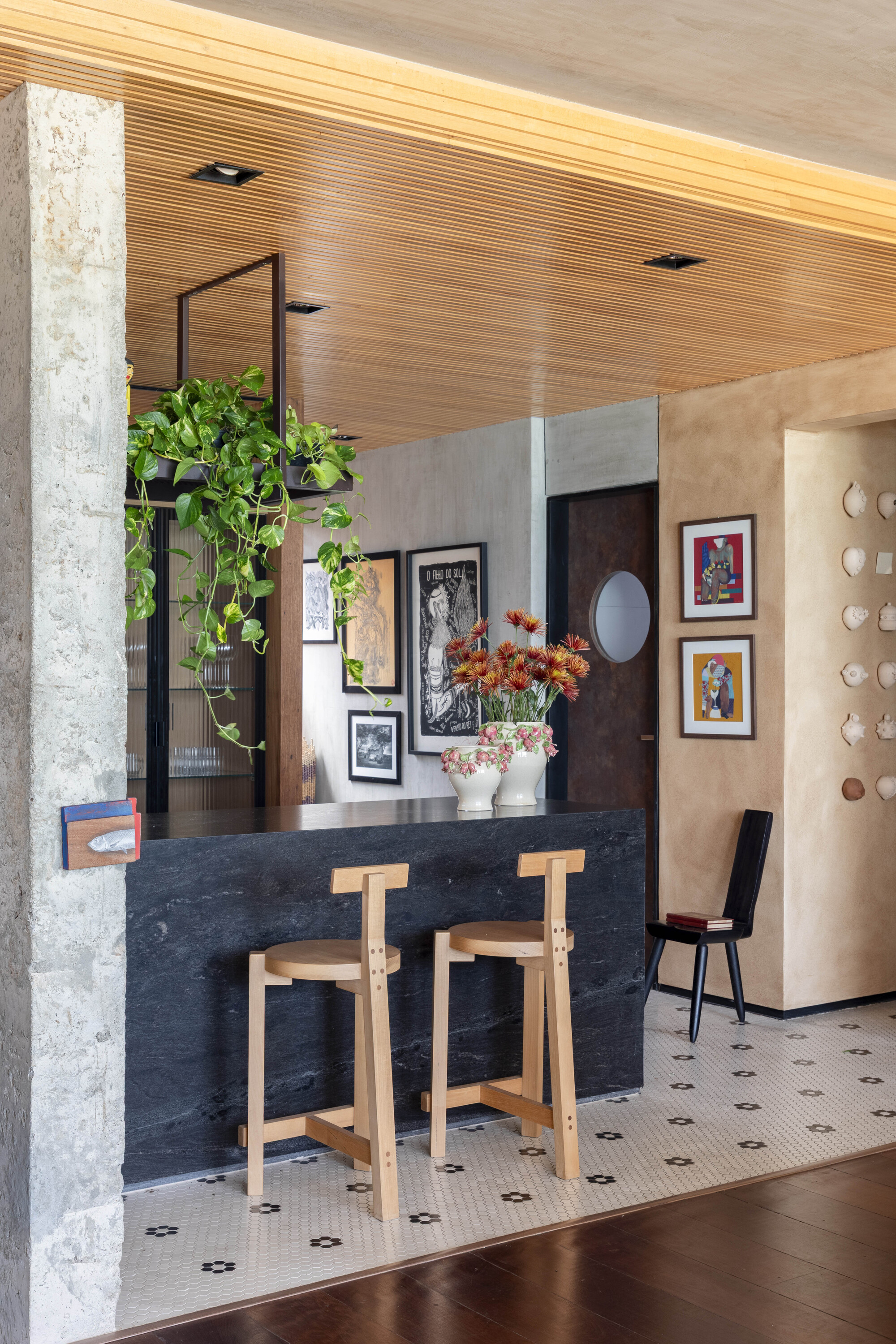 Black stone breakfast bar with two wooden stools, a hanging pothos and a slatted timber ceiling above mosaic floor tiles