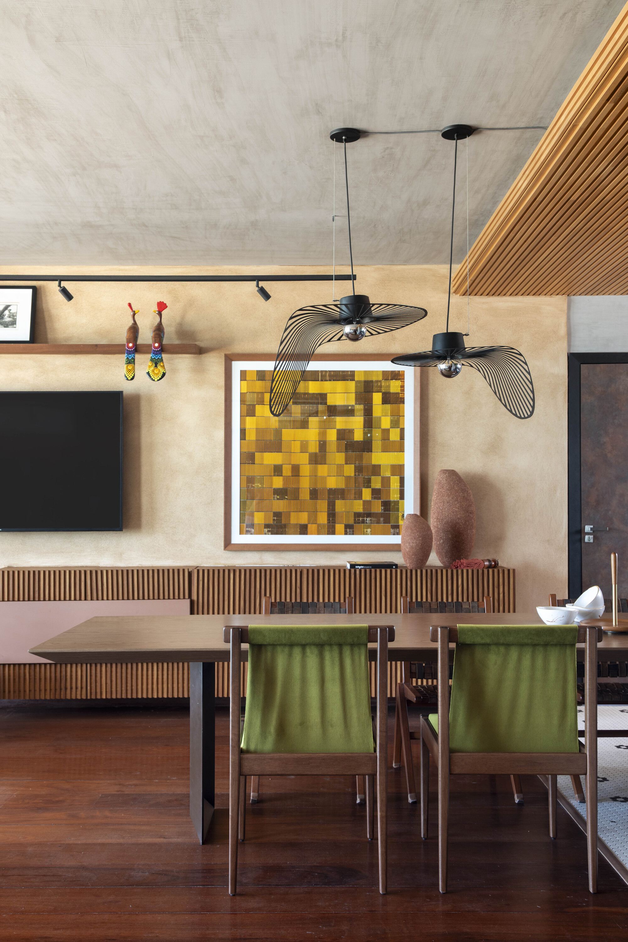 Dining area with green upholstered chairs, two wire pendant lights and a yellow mosaic artwork above a slatted wood sideboard
