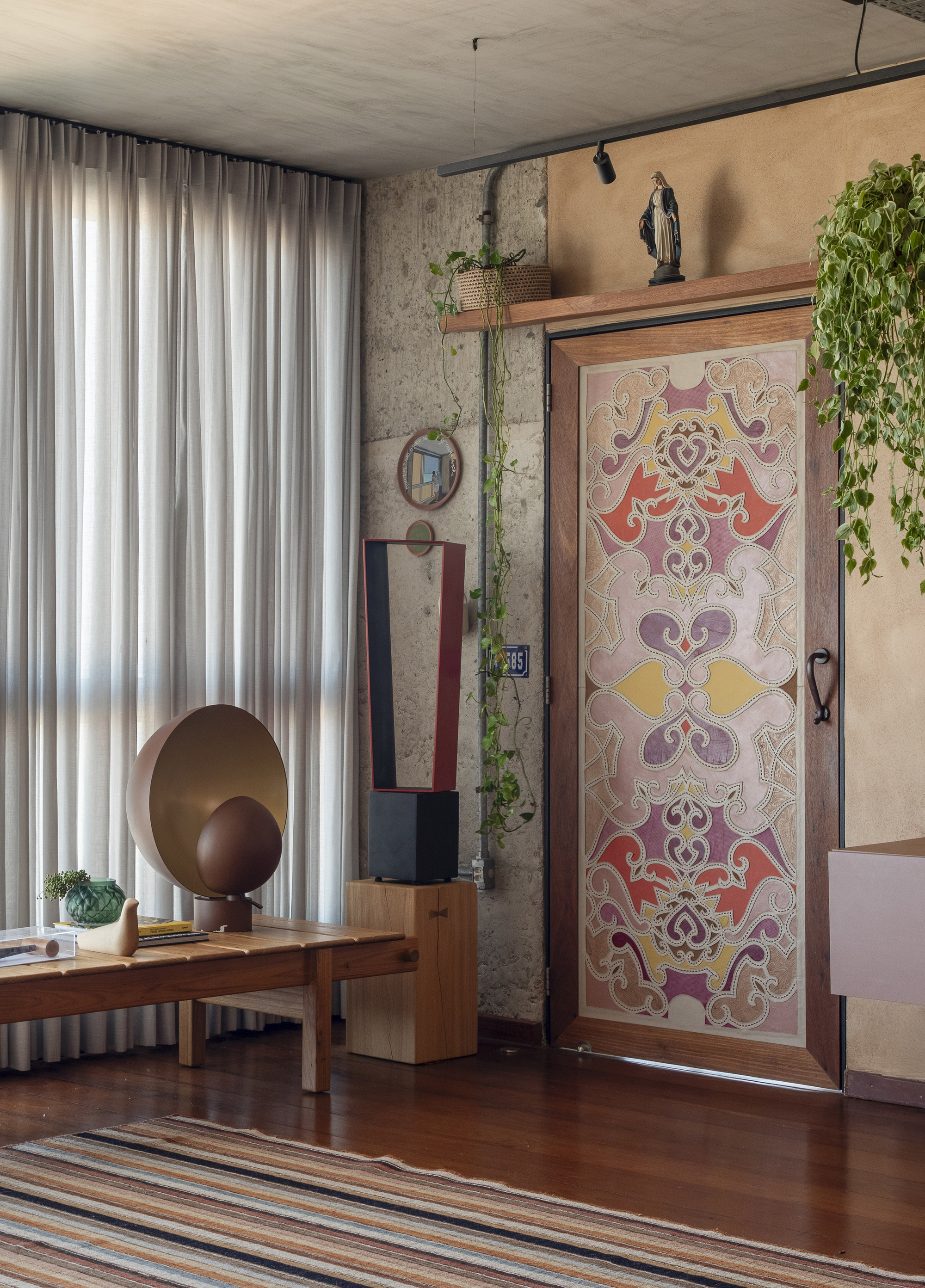 Pivot door with painted ornamental motif beside a sheer curtain, wooden bench and trailing pothos in soft daylight