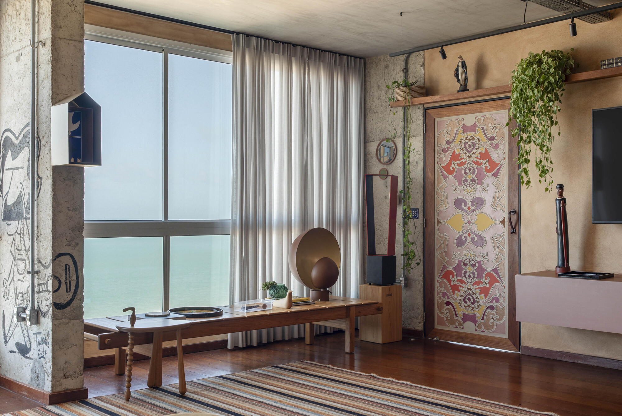 Living room corner with ornamental door, long wooden bench and graffiti-marked concrete pillar facing a hazy sea view