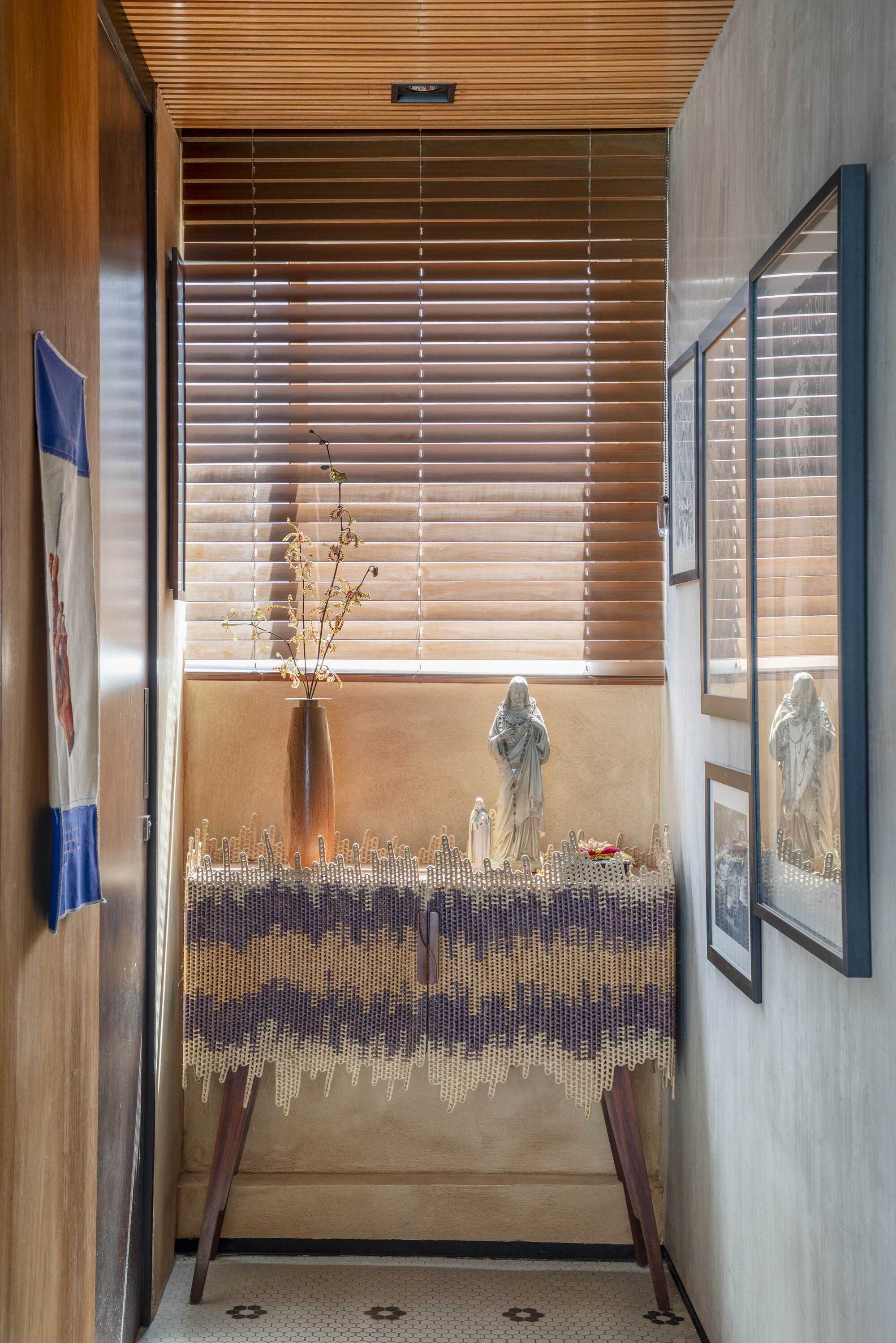 Narrow entry nook with a crocheted-edge console, devotional figurines and framed prints lit by slatted blinds