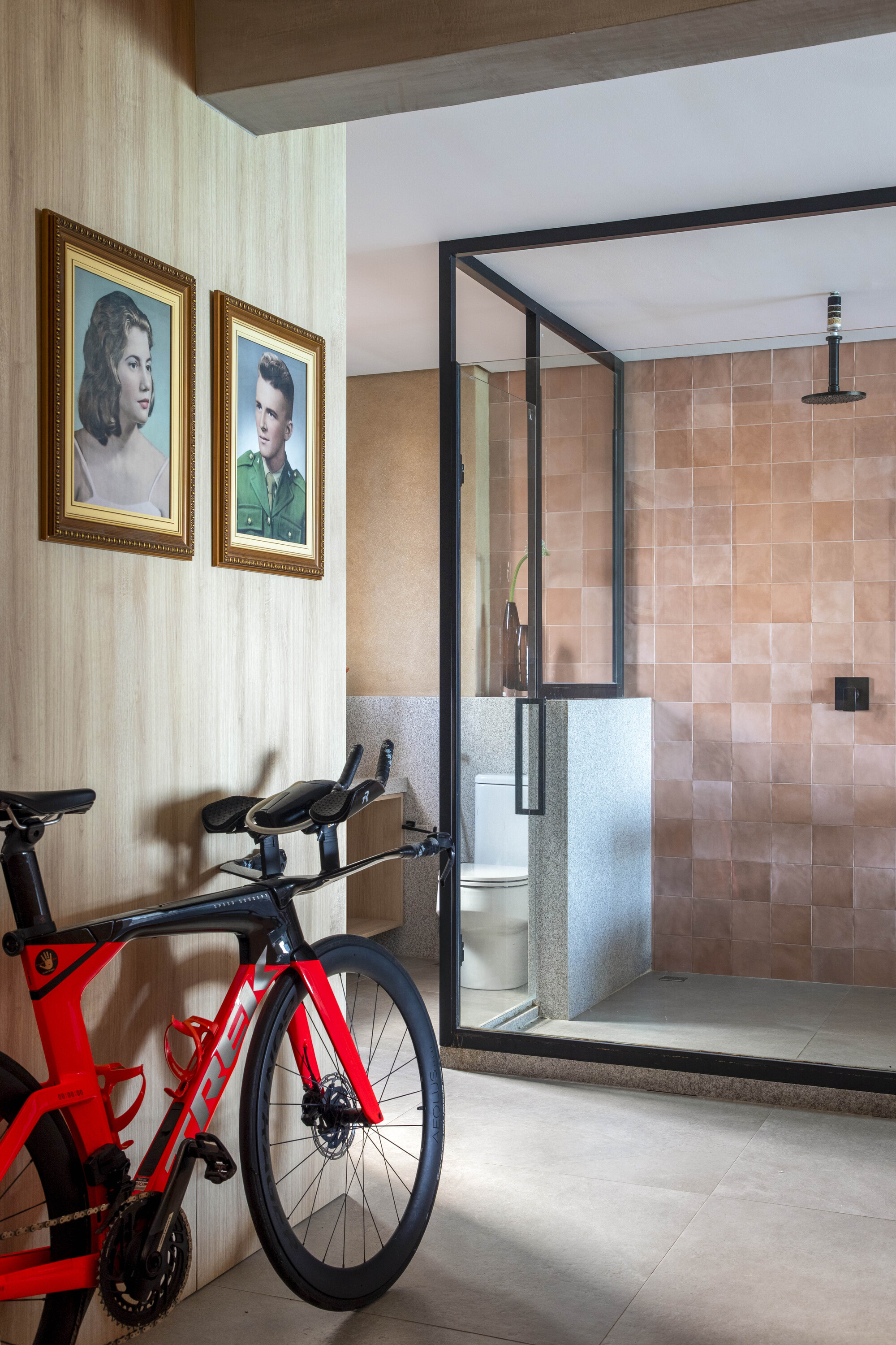 Glass-enclosed shower with pink tile walls beside a red racing bicycle and two framed portraits on a timber panel wall