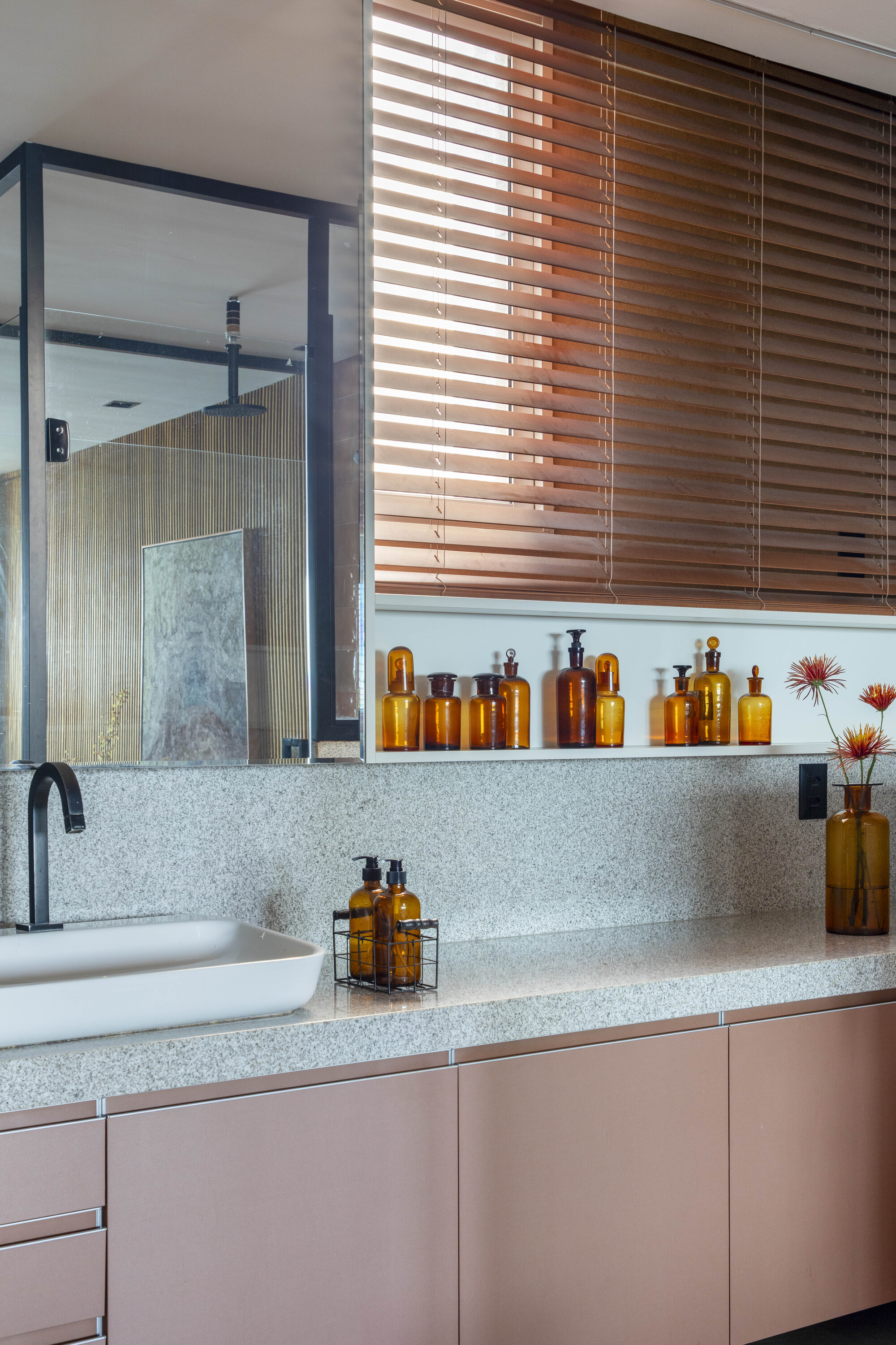 Granite vanity counter with a white basin and amber glass bottles on a shelf below half-closed wooden blinds