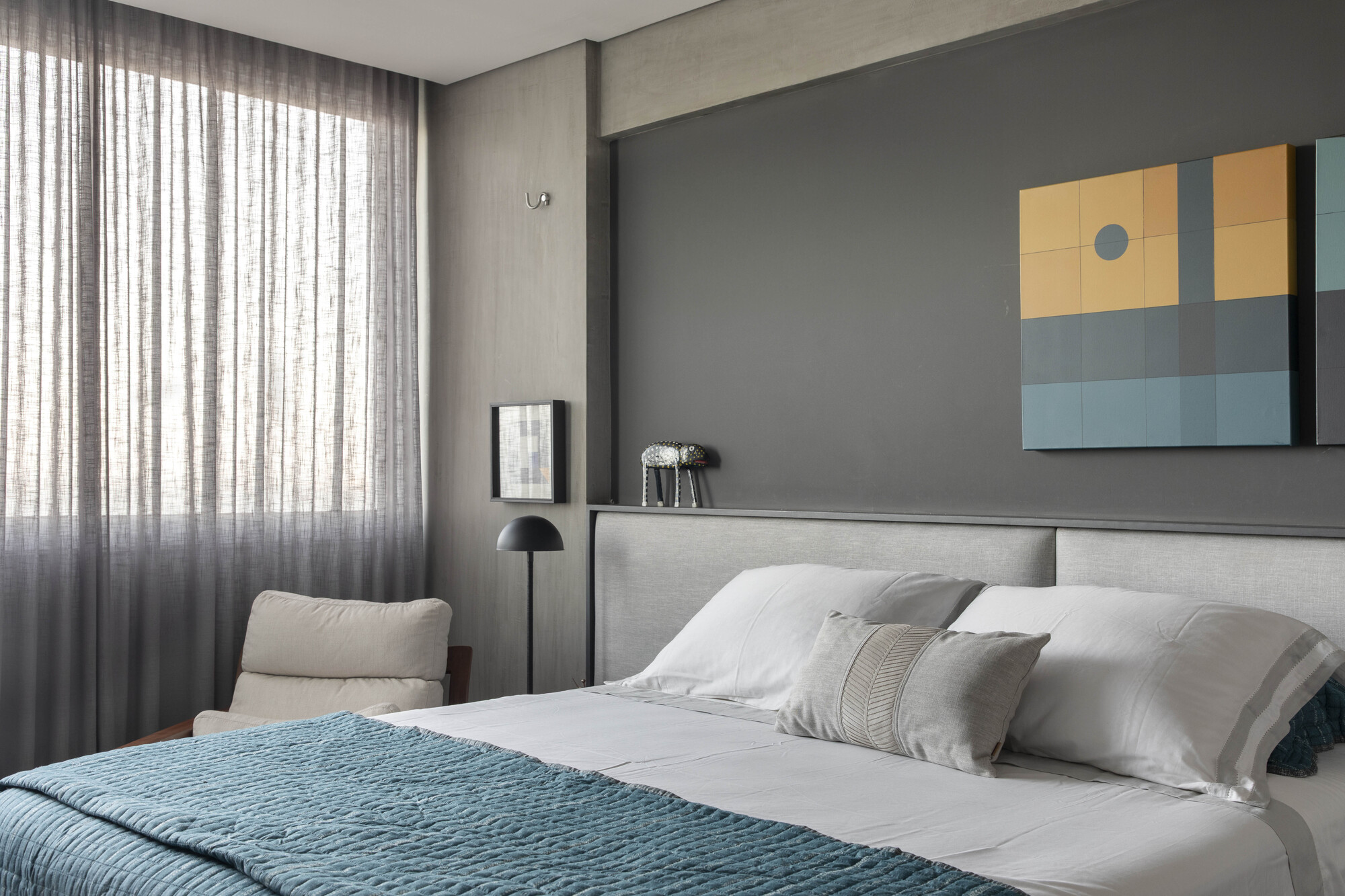 Bedroom with a grey upholstered headboard, teal quilt and sheer curtains filtering daylight beside a small armchair