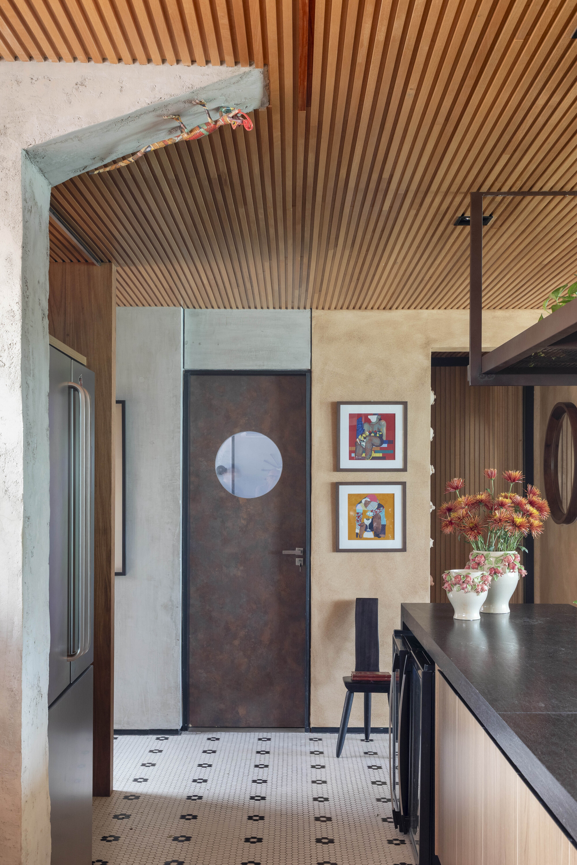 Kitchen corridor with a slatted timber ceiling, a round-windowed rusted metal door and hex-patterned floor tile beside a dark countertop