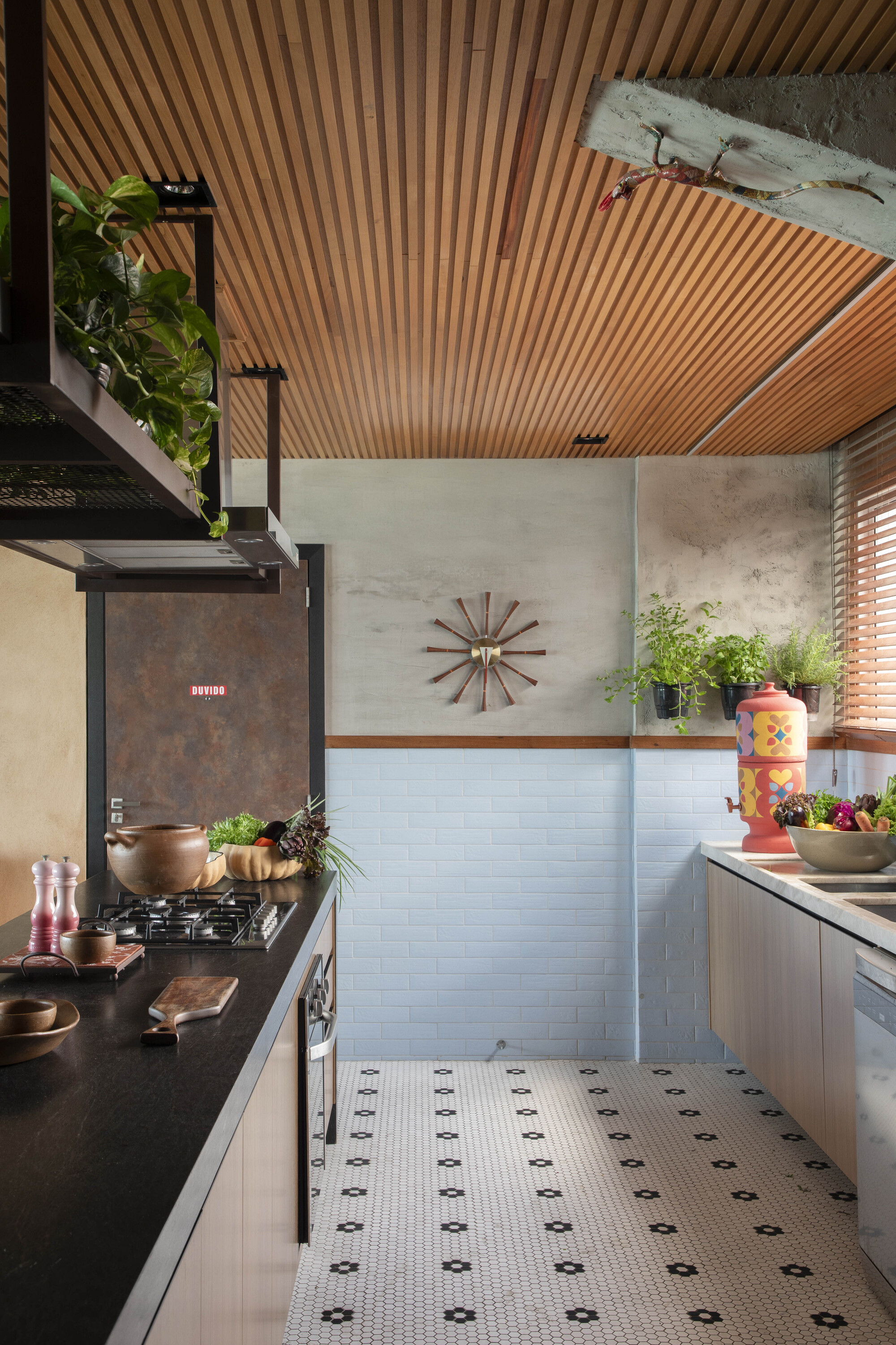 Galley kitchen with slatted timber ceiling, pale blue brick wall and hexagonal floor tile, with potted herbs catching window light