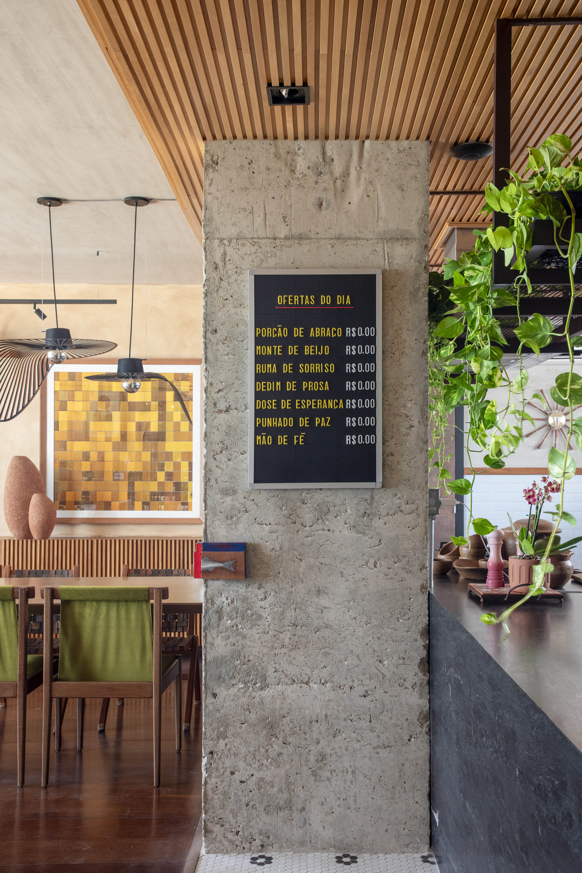 Raw concrete column carrying a chalkboard menu, with trailing pothos hanging beside the dark counter and dining chairs beyond
