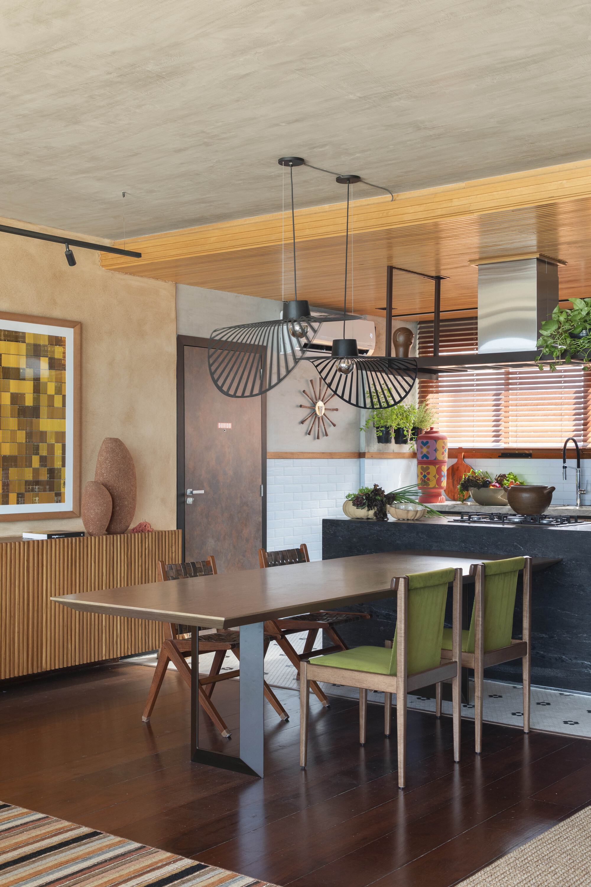 Dining table with green upholstered chairs under woven pendant lamps, with the kitchen island and timber ceiling behind in warm light
