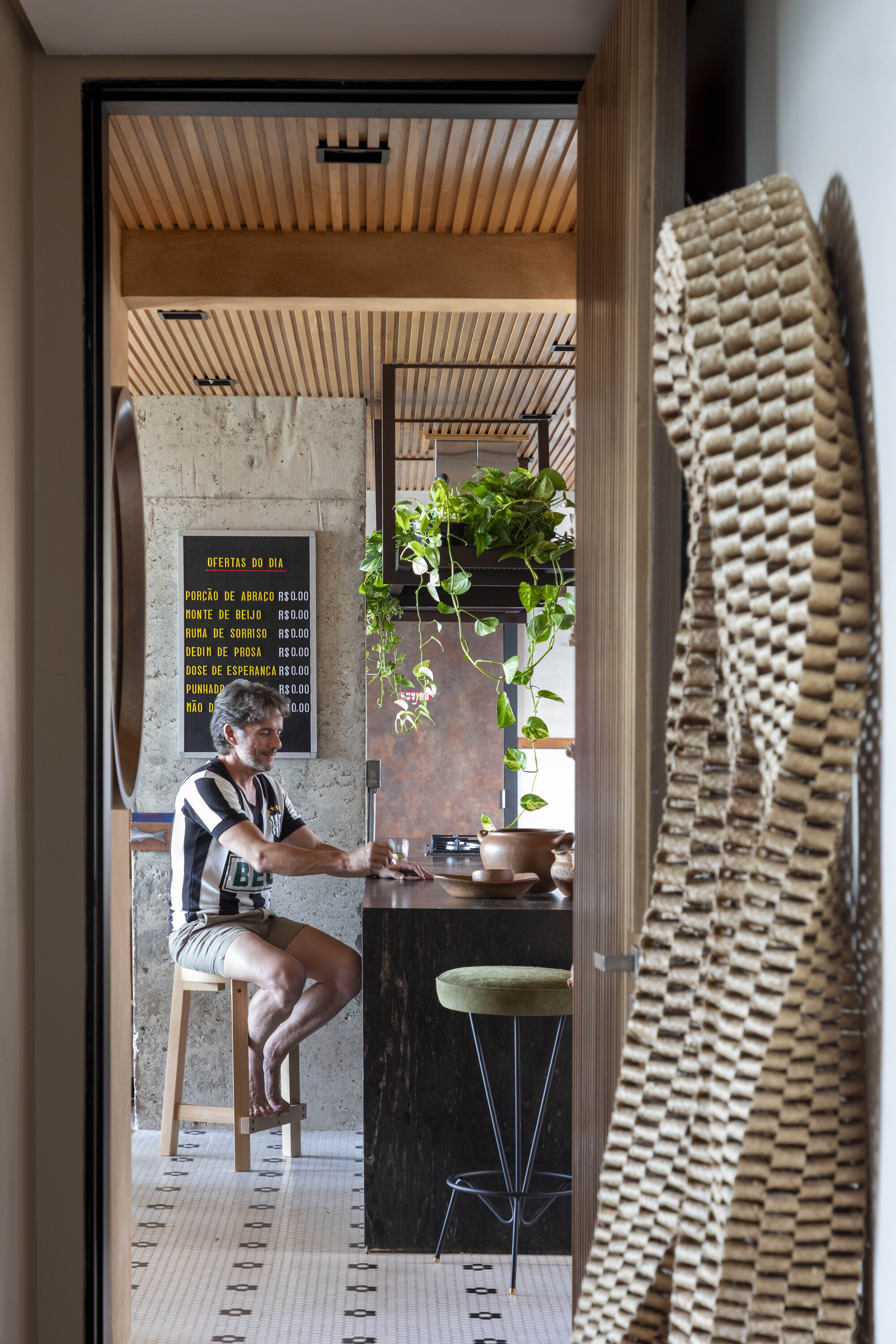 Doorway view of a man seated at the kitchen counter beneath a chalkboard menu, with a woven textile hanging in the foreground