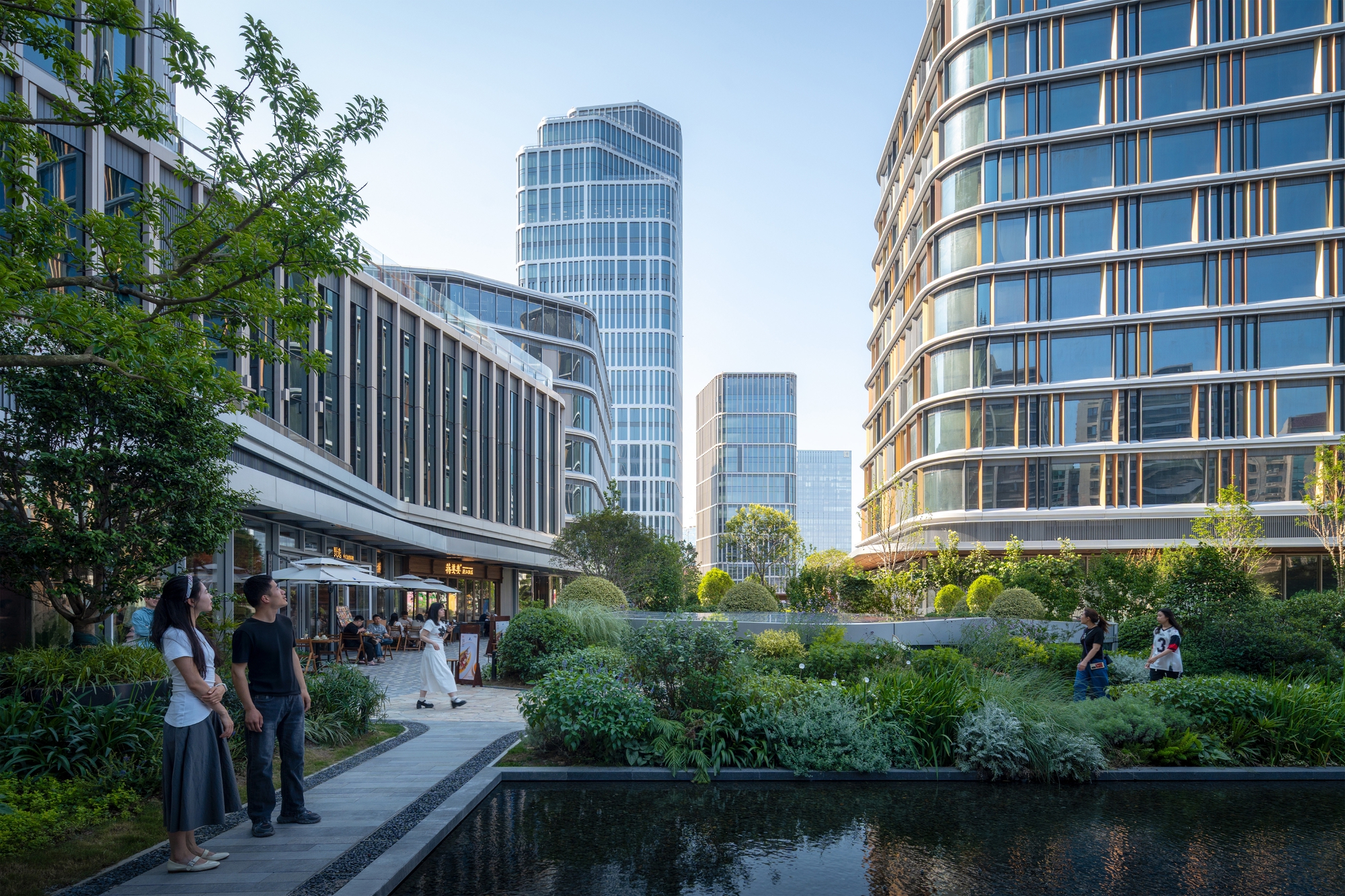 Garden courtyard with a reflecting pool and a stone path, with curved glass towers rising behind visitors in late afternoon sun