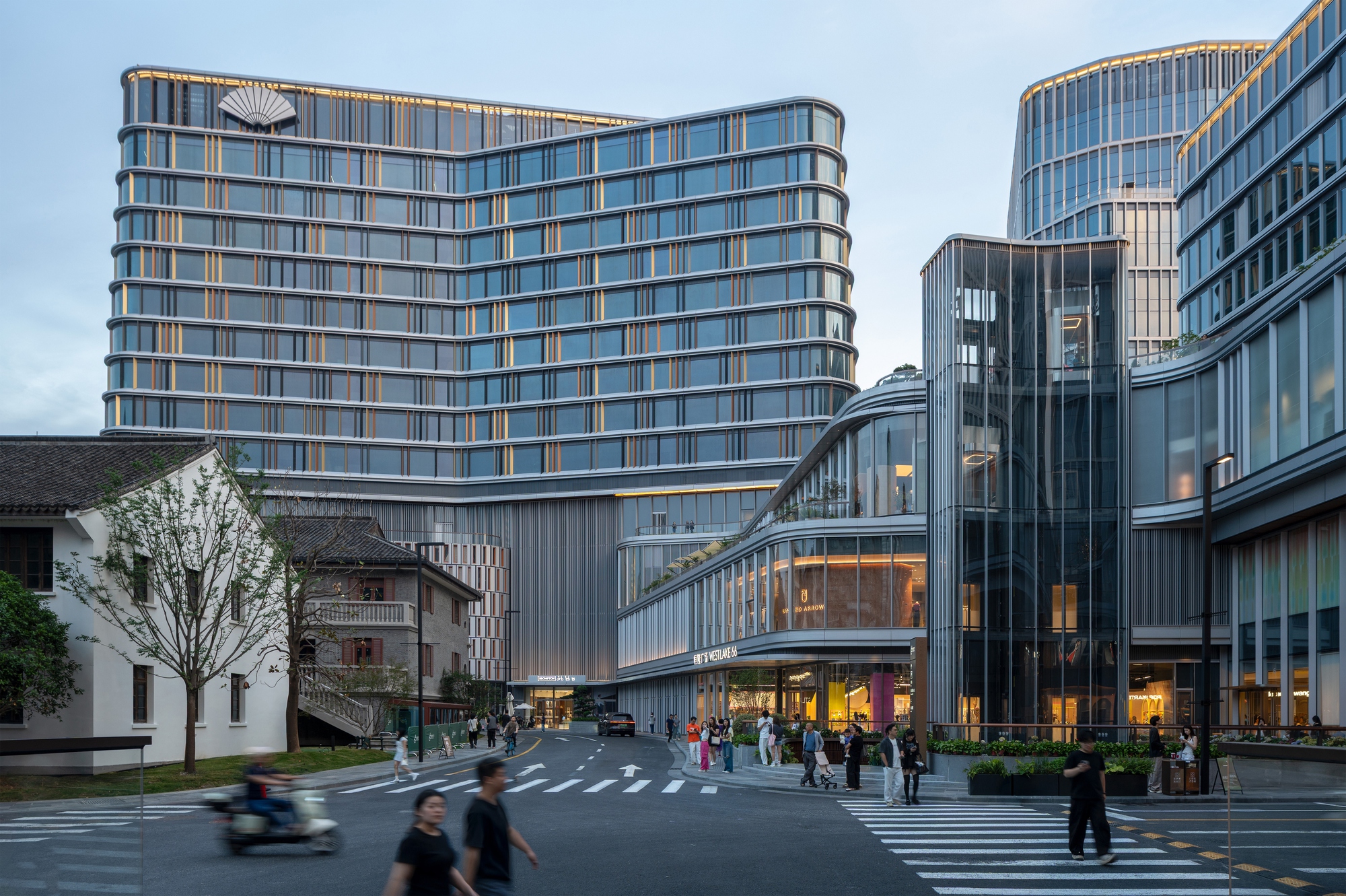 Street corner with a curved, banded glass tower and a glazed lobby volume beside a low old tiled-roof house at dusk