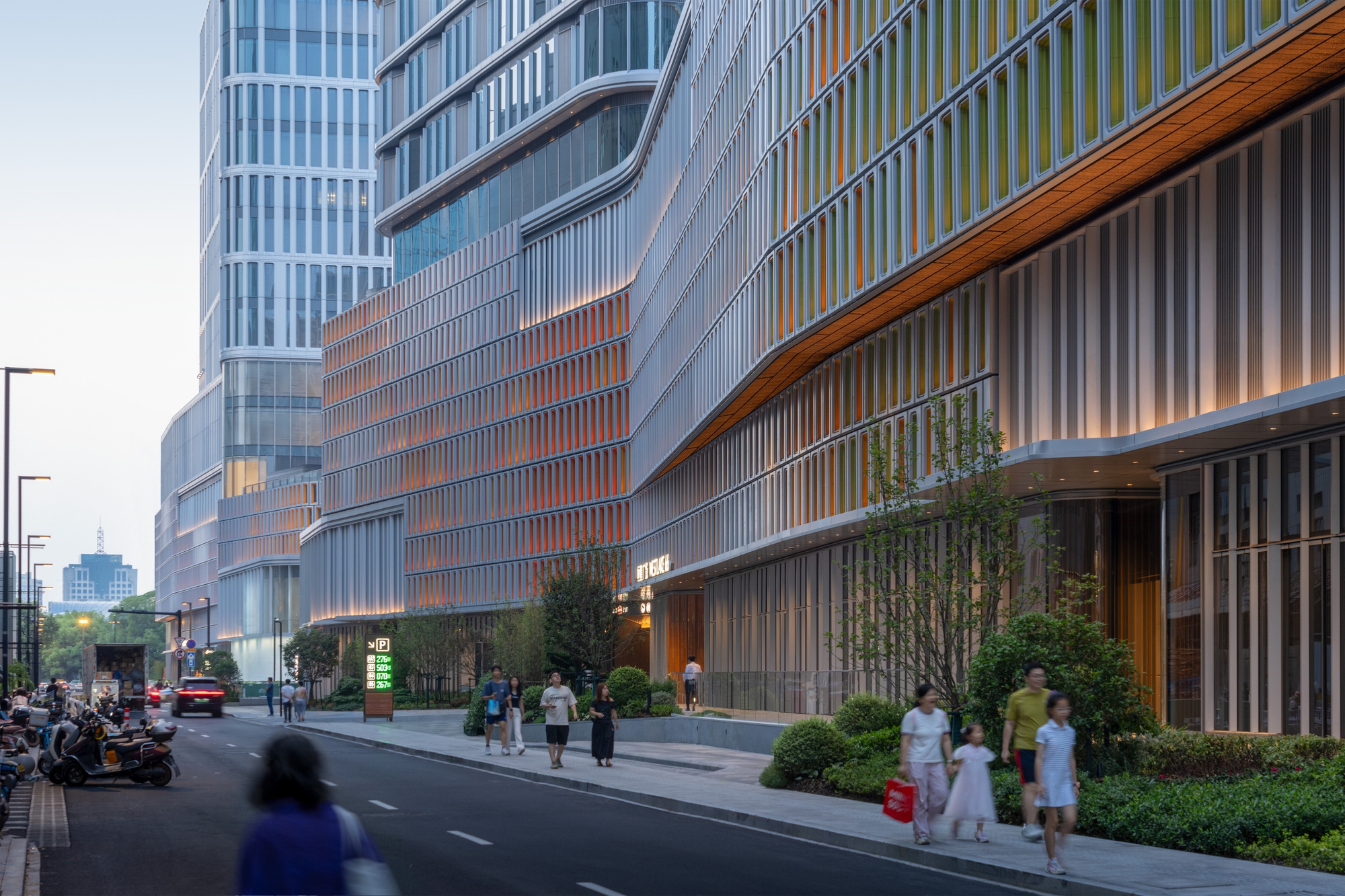 Street-side facade of vertical fins glowing orange with a cantilevered timber soffit, with families walking the pavement at dusk