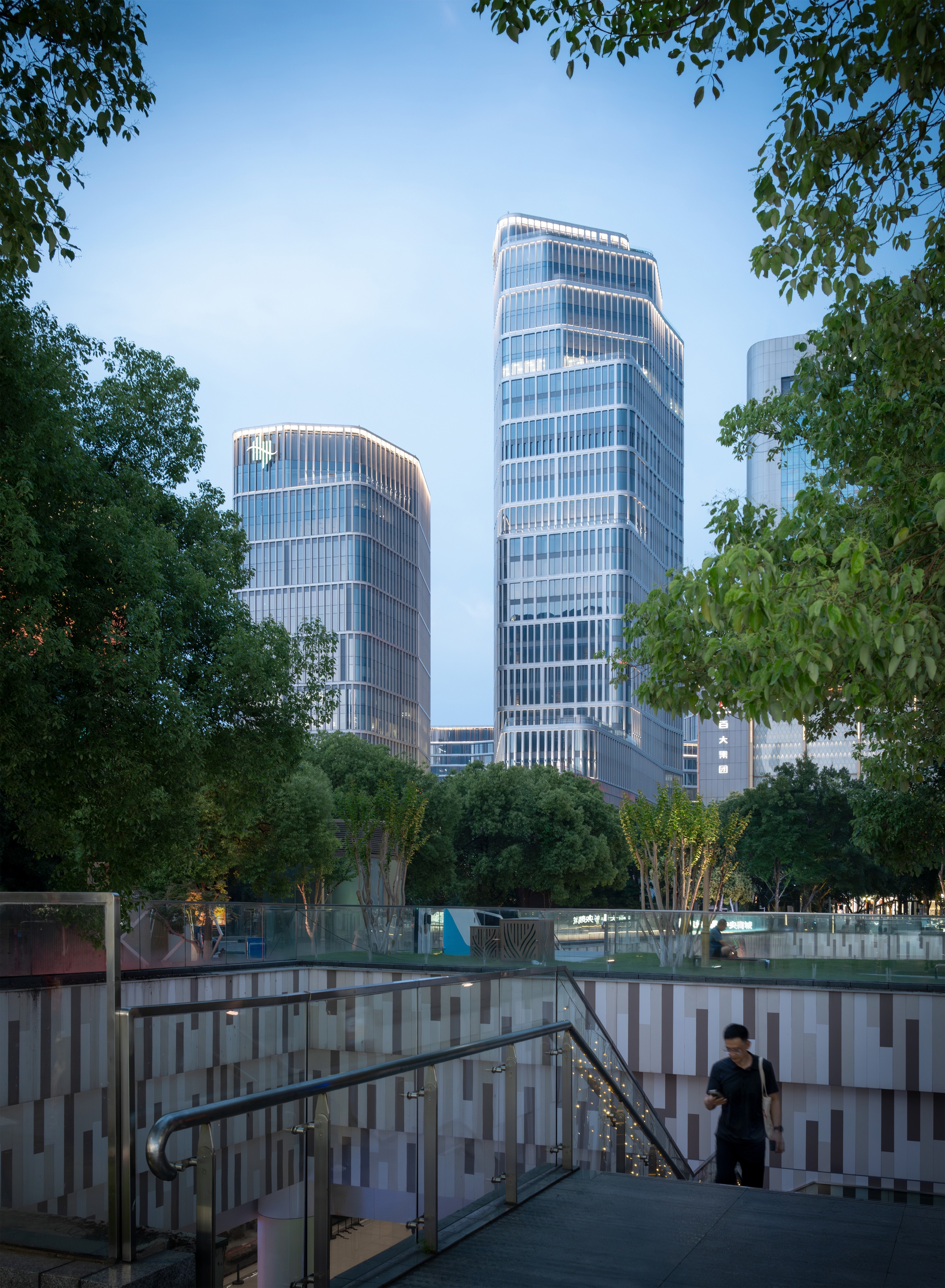 Glass-balustraded stair with a patterned stone wall, leading toward two glass towers framed by tree canopy under a blue evening sky