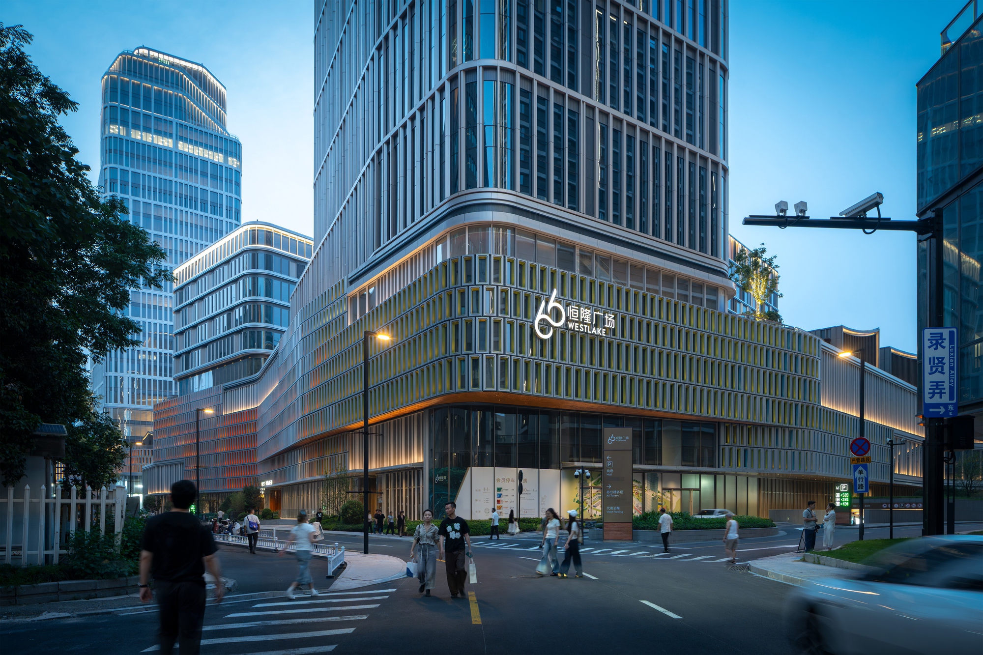 Podium facade with green vertical fins and a rooftop sign, below stepped glass towers, with pedestrians crossing the street at blue hour