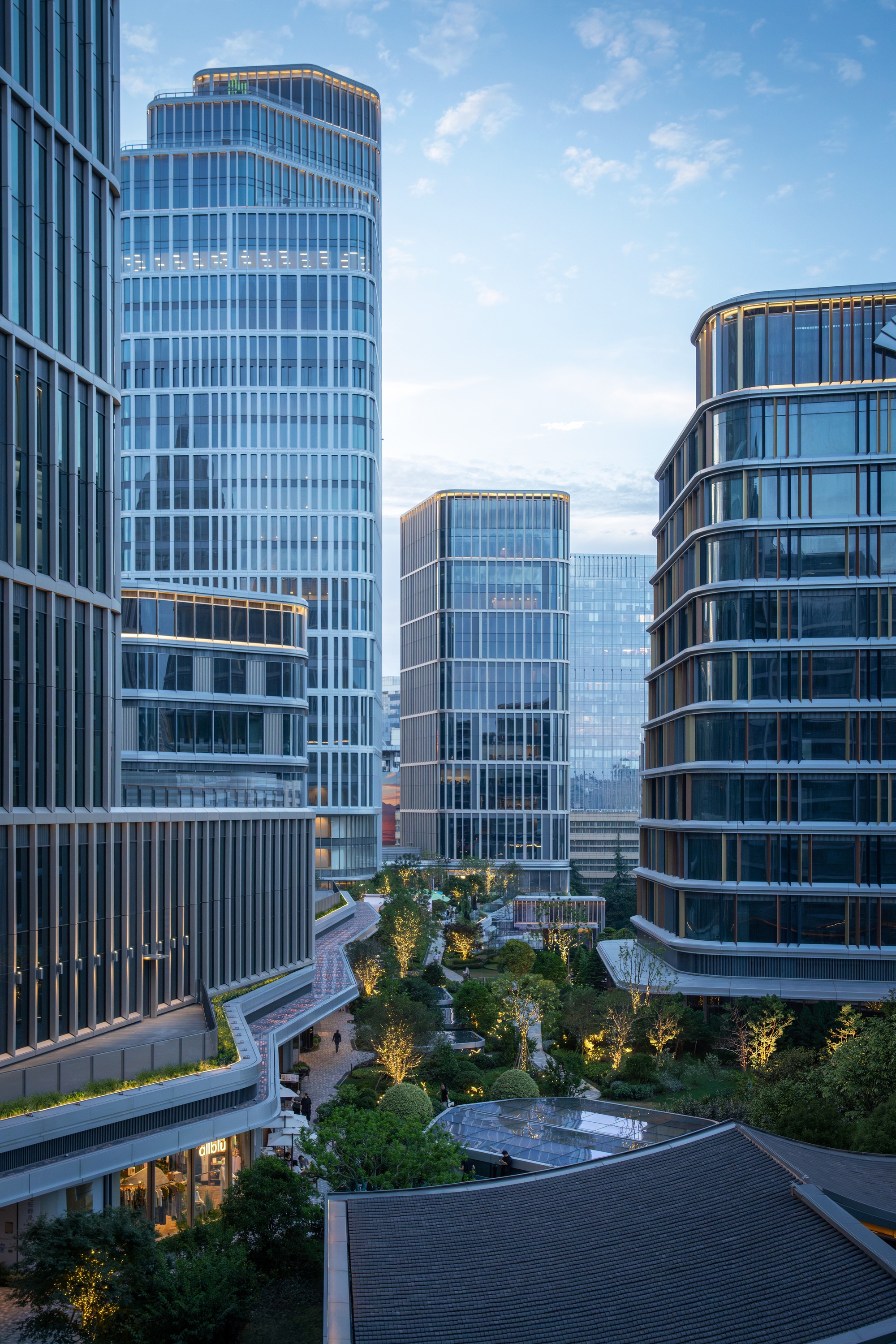 Glass towers rising above a landscaped courtyard with lit trees and a green roof terrace at dusk