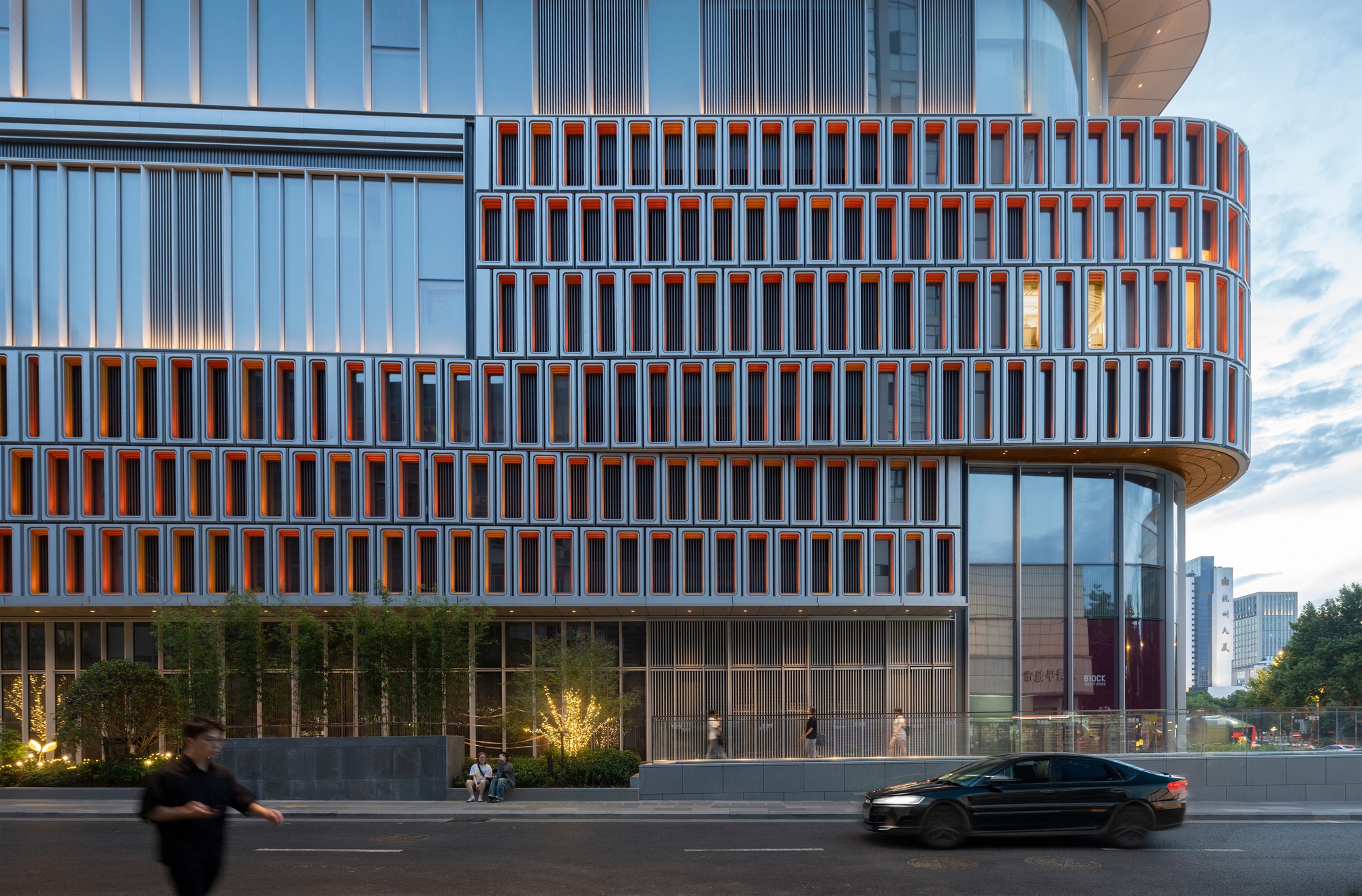 Street facade with orange-lined window frames over a bamboo planting strip, a passing car and pedestrians in evening light