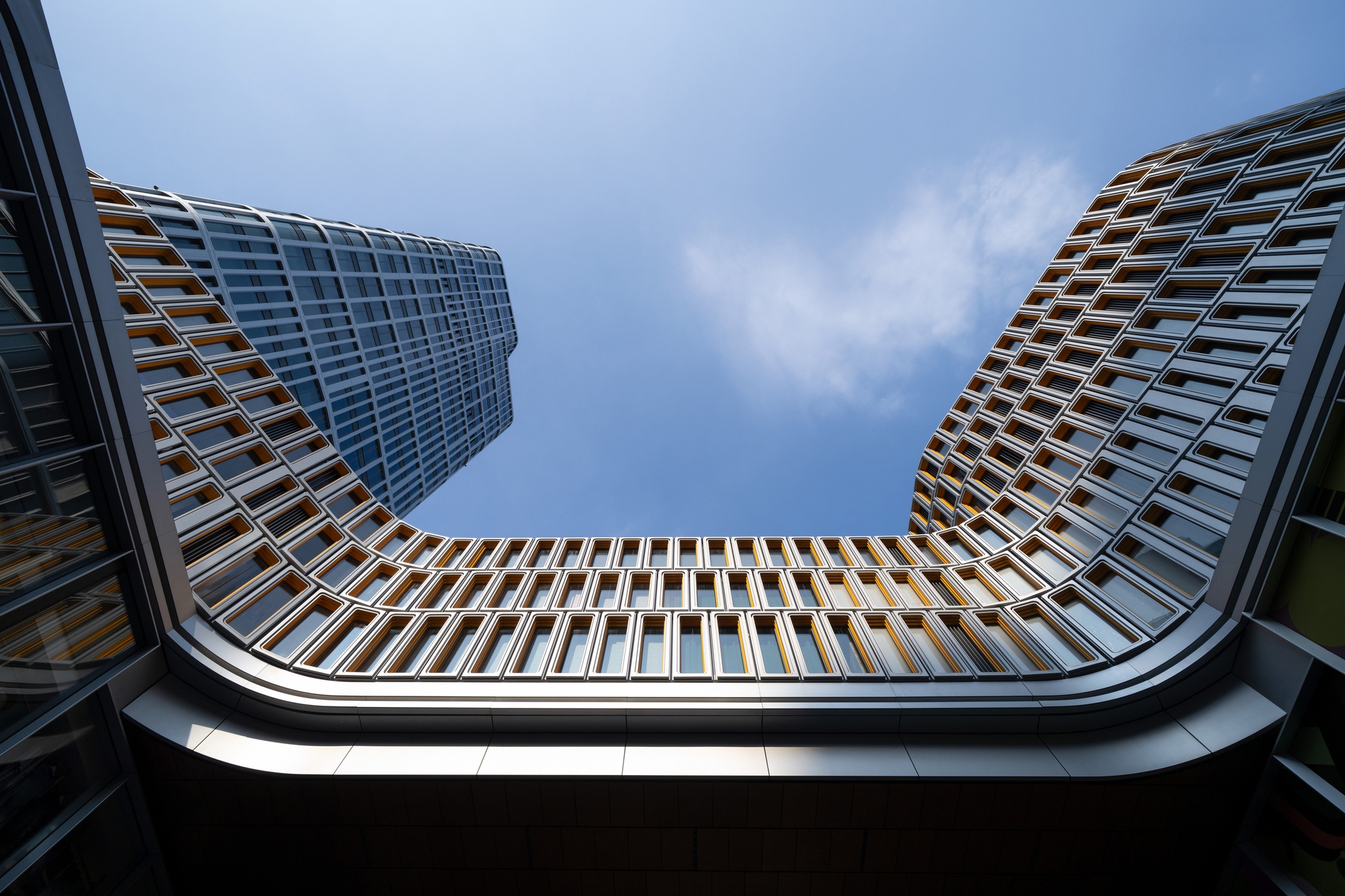 Upward view through a curved opening framed by gridded window facades against a blue sky with a thin cloud