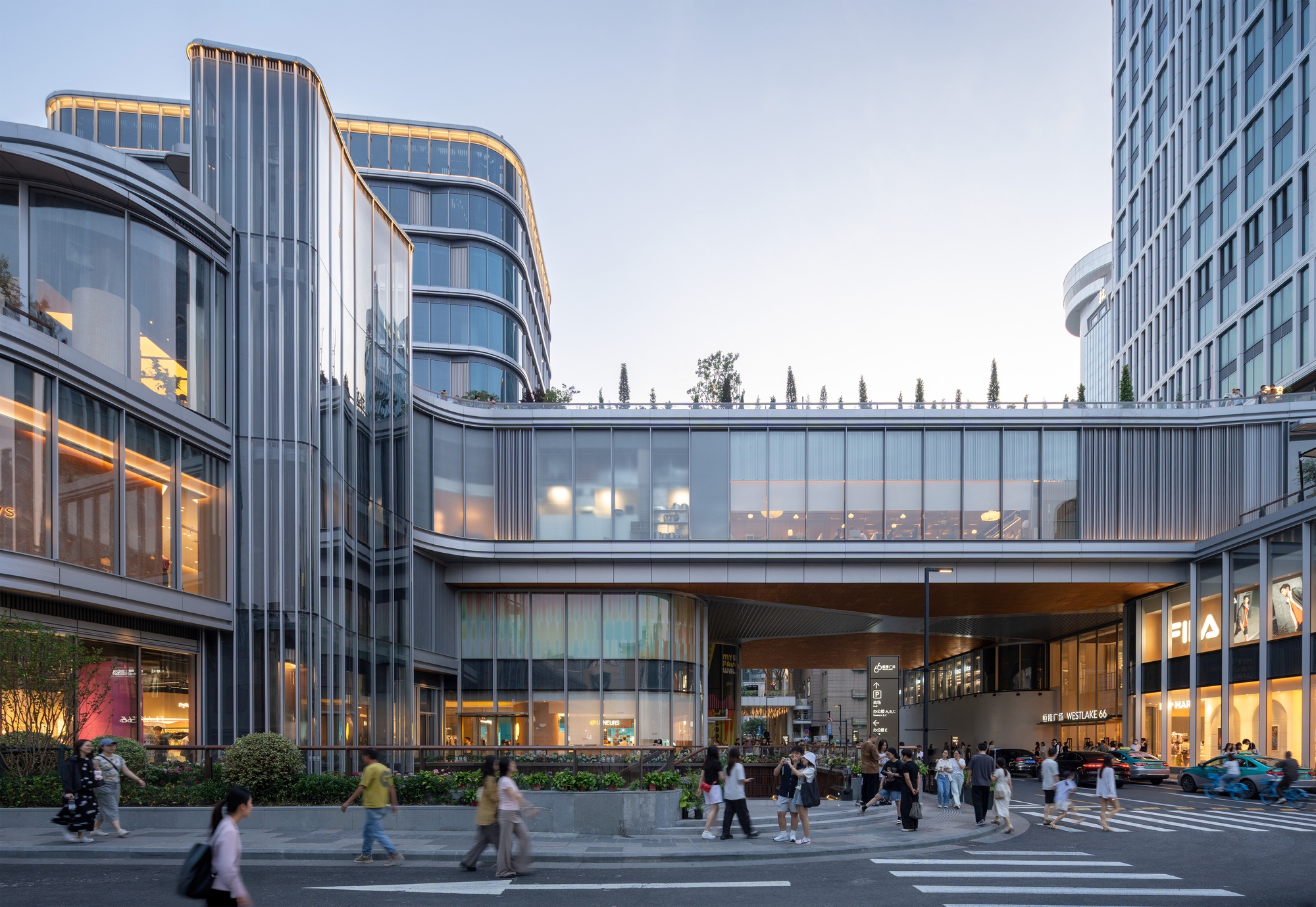 Pedestrian crossing in front of a glazed sky bridge linking two curved podium volumes at dusk
