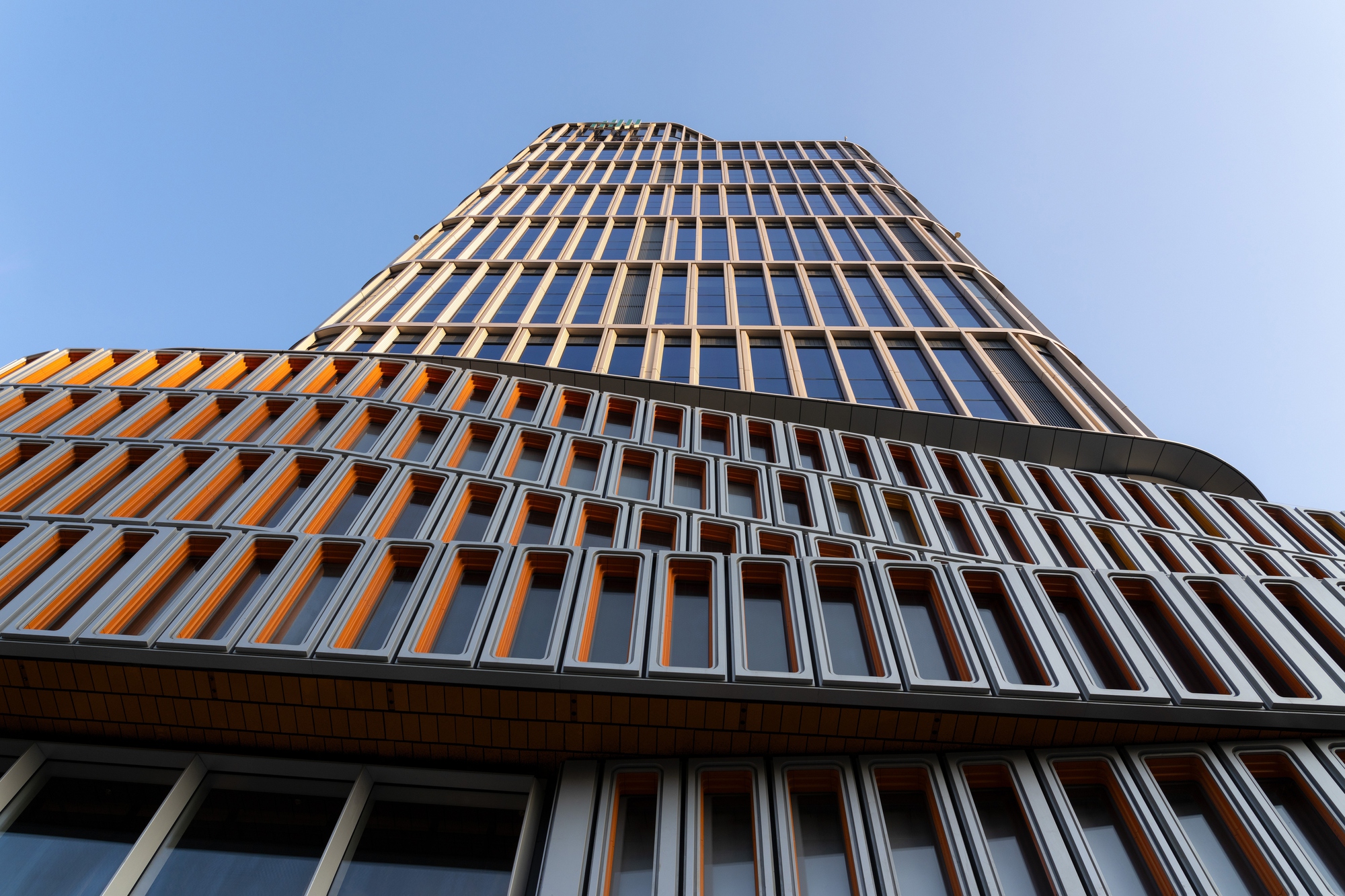 Low-angle view of a tower stepping back above rows of orange-trimmed window frames under a clear blue sky