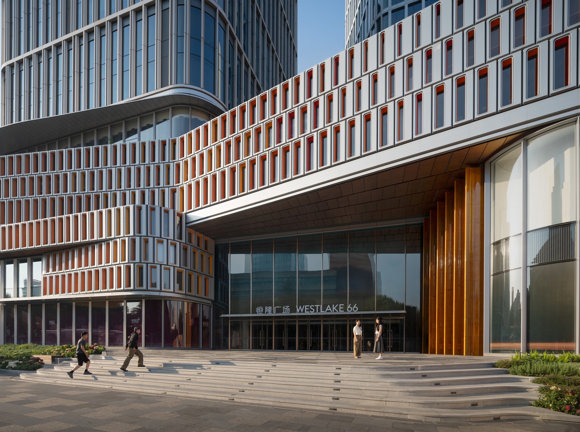 Entrance canopy with timber-clad fins and a facade of orange-lined window boxes above wide stone steps, with pedestrians in late sun