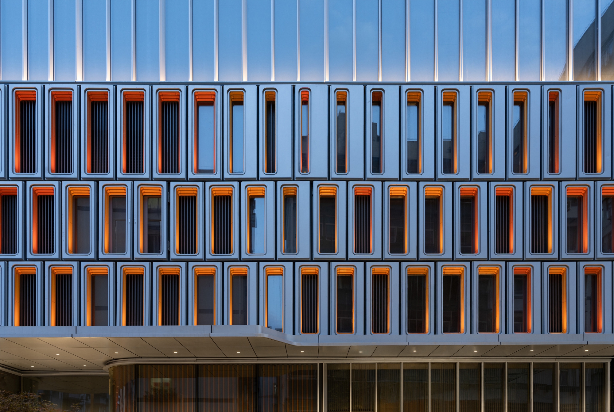 Frontal view of the aluminium window-box facade with orange-lit recesses and louvres, glowing at dusk above a curved canopy