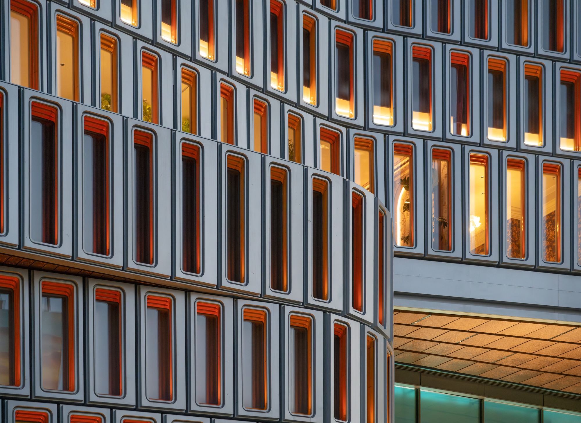 Close-up of curved facade panels with orange-edged windows, lit warmly from inside at dusk beside a timber-lined soffit
