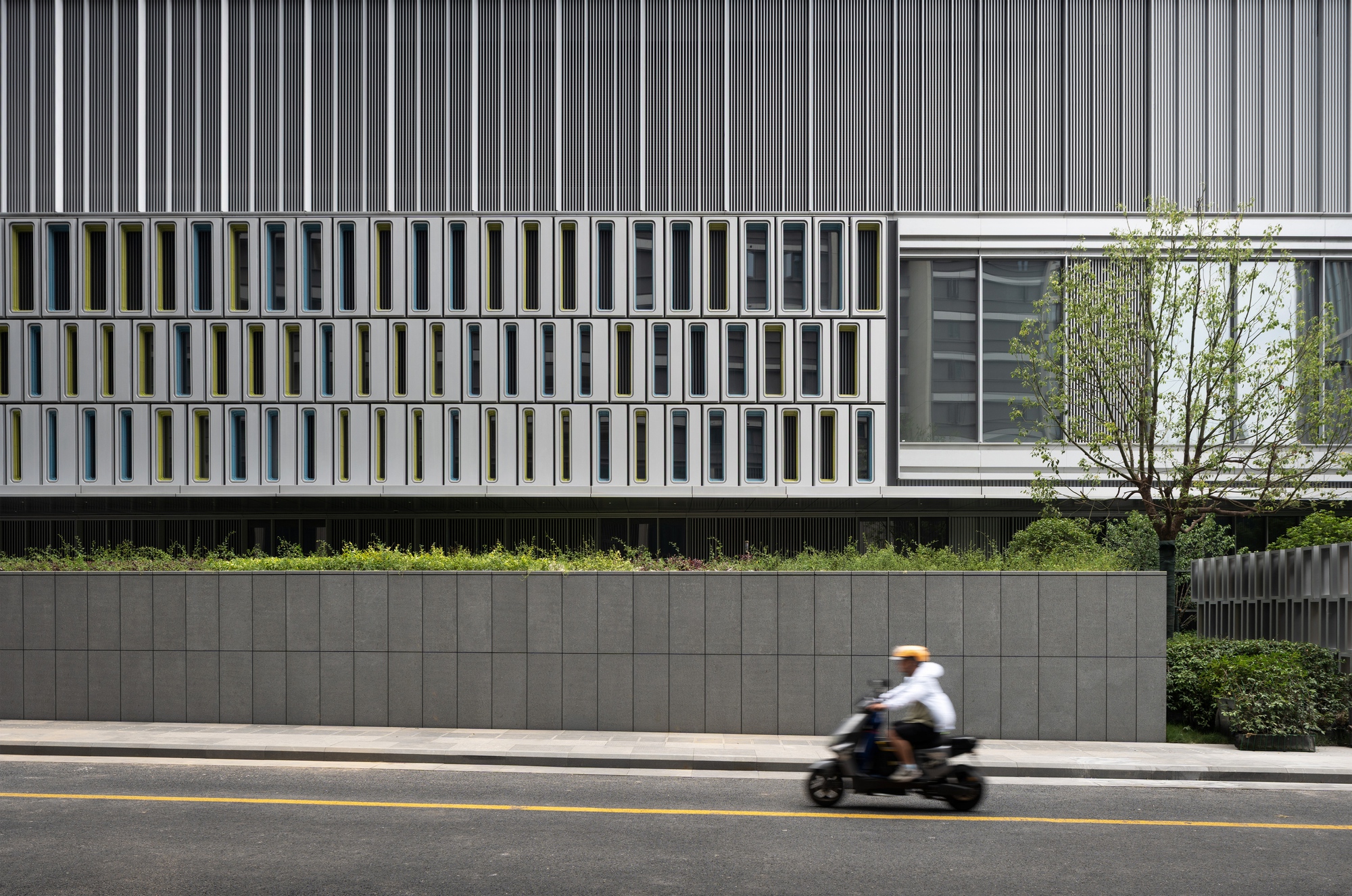 Street-level view of a facade with small yellow and blue window frames above a stone wall, with a scooter rider passing