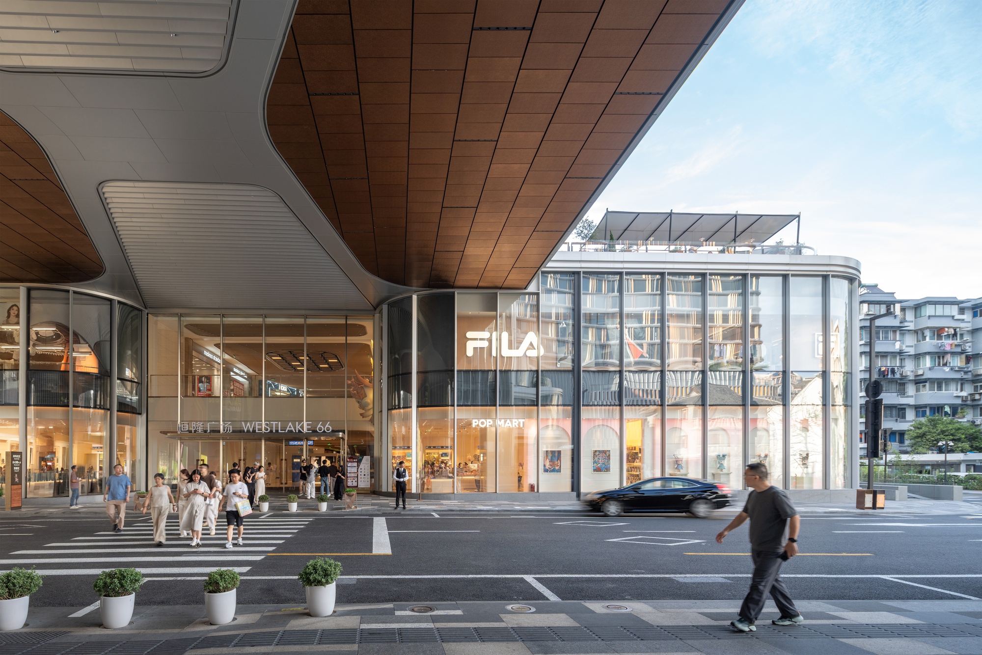 Timber-lined canopy overhanging a glazed retail corner at a street crossing, with pedestrians and a passing car in daylight