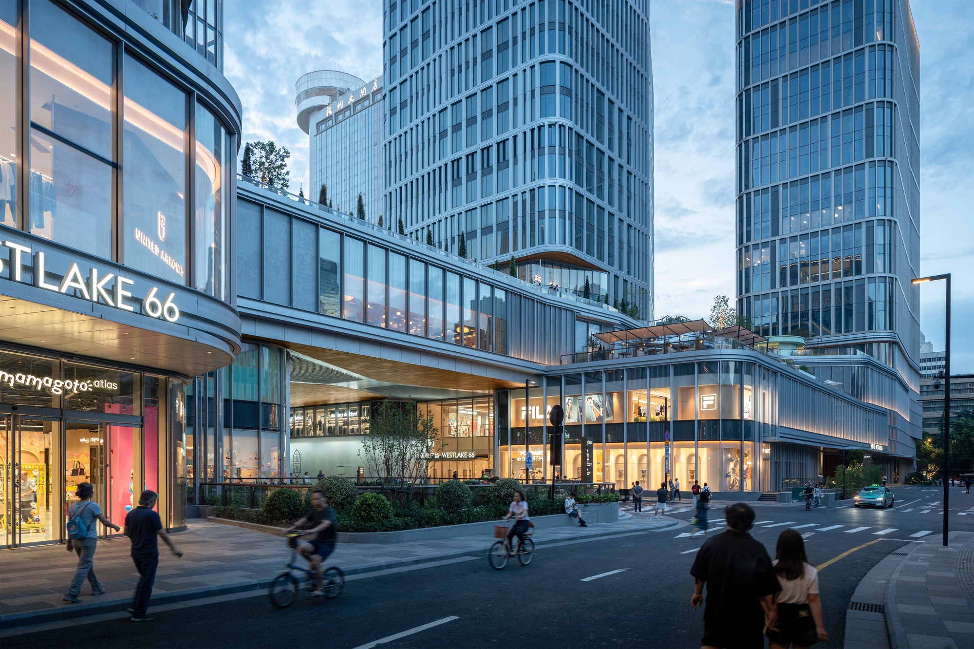Street corner of a glass-fronted retail podium below tall towers, with cyclists and pedestrians passing at dusk